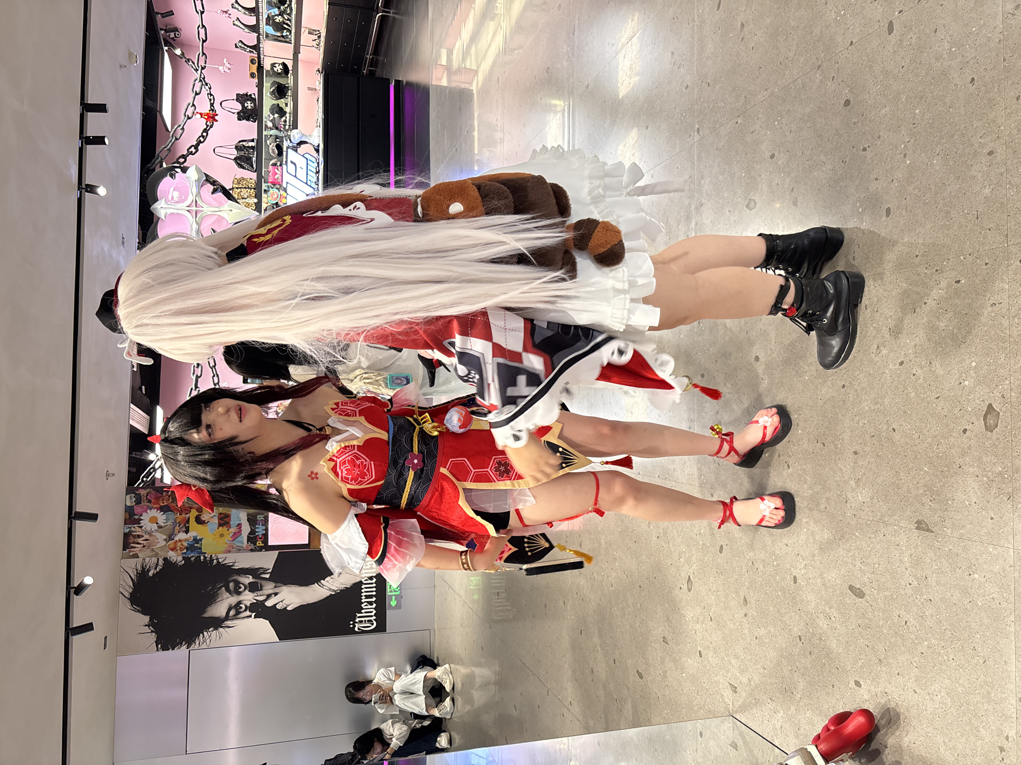 Here is a detailed description of the image:
**Scene:**
The image captures two individuals in cosplay outfits. The setting appears to be inside a retail space or a commercial area with fashion displays, posters, and other shoppers in the background. The floor is gray and seems to be made of stone or concrete-like material.
**People:**
Two people are captured in the frame, both dressed in elaborate cosplay costumes.
* The first person has black hair with red accents and is wearing a red and gold costume with matching red sandals.
* The second person is wearing a costume that incorporates a long white wig, a white and red dress, and black boots. They also have a brown plush toy attached to their outfit.
**Objects:**
* Fashion displays with various items such as bags and accessories are visible in the background.
* Posters or advertisements are displayed on the wall.
* A pair of red boxing gloves are located on the floor in the foreground.
**Actions/Interactions:**
The cosplayers appear to be posing or standing next to each other. There isn't much interaction captured in the image other than the two people being close together.
**Environment:**
The environment seems to be a commercial space likely in a shopping center. There are signs of active retail or display setups. The scene is indoors, illuminated by indoor lighting.