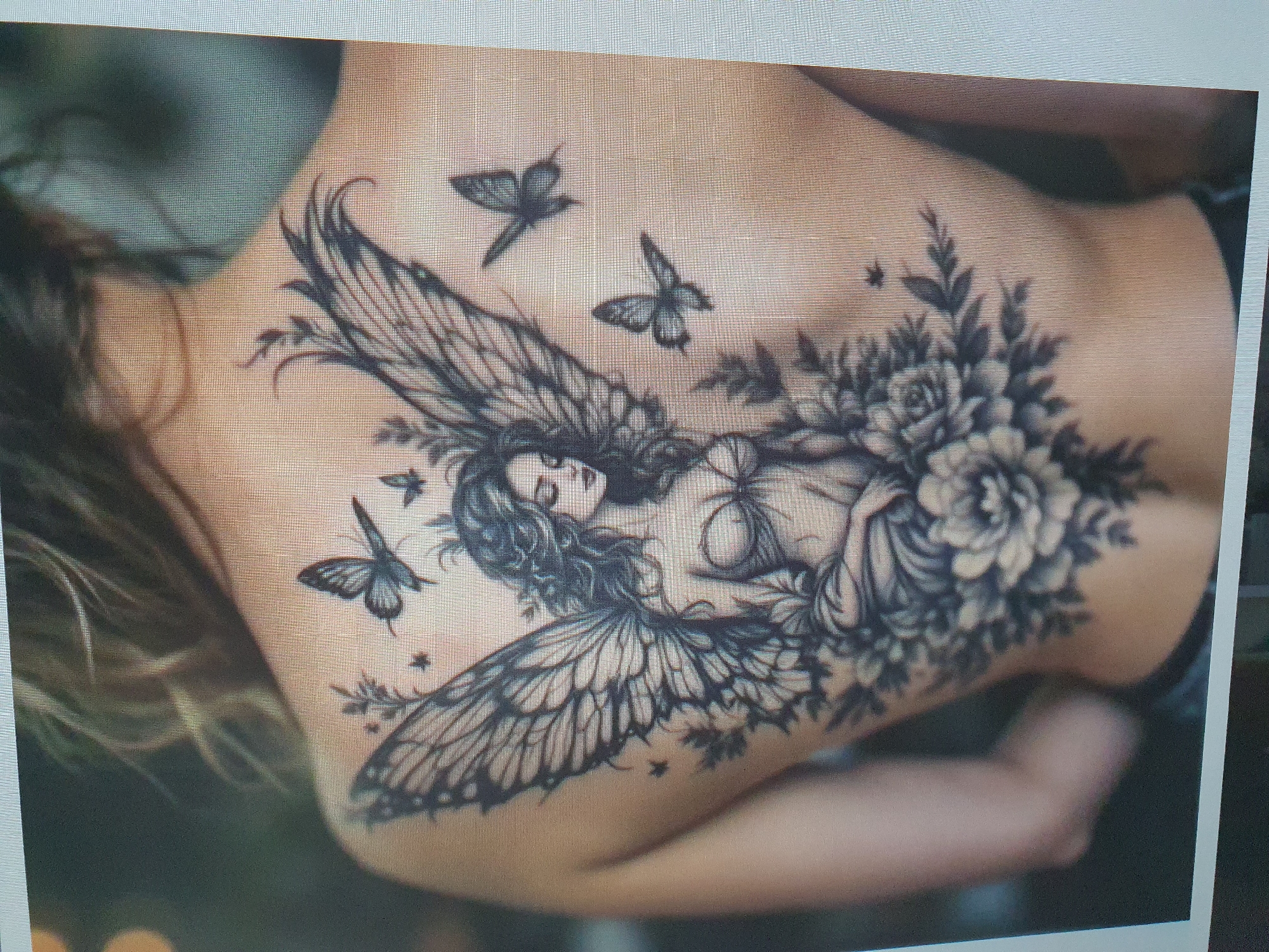 The media file shows a close-up of a tattoo on a person's back. The tattoo is a detailed black and grey design featuring a woman with butterfly wings, surrounded by flowers and smaller butterflies. The woman's face is visible, with her eyes closed and hair flowing. The tattoo covers a significant portion of the back, extending from the shoulders down towards the lower back. The setting appears to be indoors, with a soft, diffused lighting. There is no visible text in the image. The overall mood conveyed by the tattoo is one of ethereal beauty and fantasy.