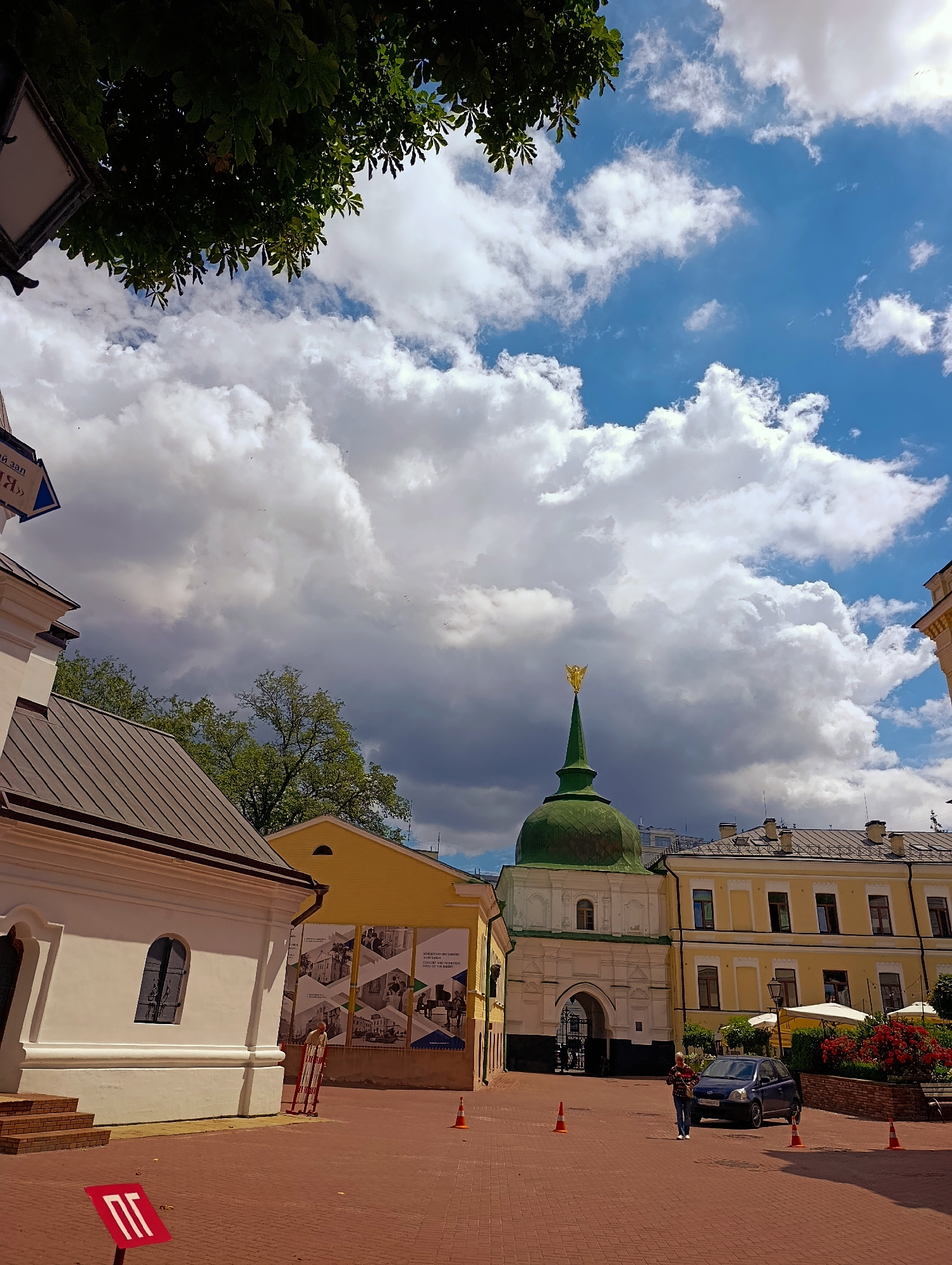 The image shows the Kyivo-Pecherska Lavra in Kyiv, Ukraine, on a sunny day with dramatic clouds. In the foreground, a cobblestone courtyard is visible, with a white building to the left and a yellow building with informational posters to the right. A red sign with white Cyrillic letters is planted in the courtyard. In the mid-ground, a green dome building with a golden angel on top is prominent. To the right of the dome building is a long, yellow residential building with arched windows. A dark blue car is parked in front of this building. A woman in casual clothing stands near the car, facing away from the camera. Several orange traffic cones are placed on the ground in the courtyard. Lush green trees and red flowers are visible behind the buildings. The sky is a mix of bright blue and dark, heavy clouds, suggesting the possibility of rain. The overall scene depicts a historic religious complex with elements of urban life.