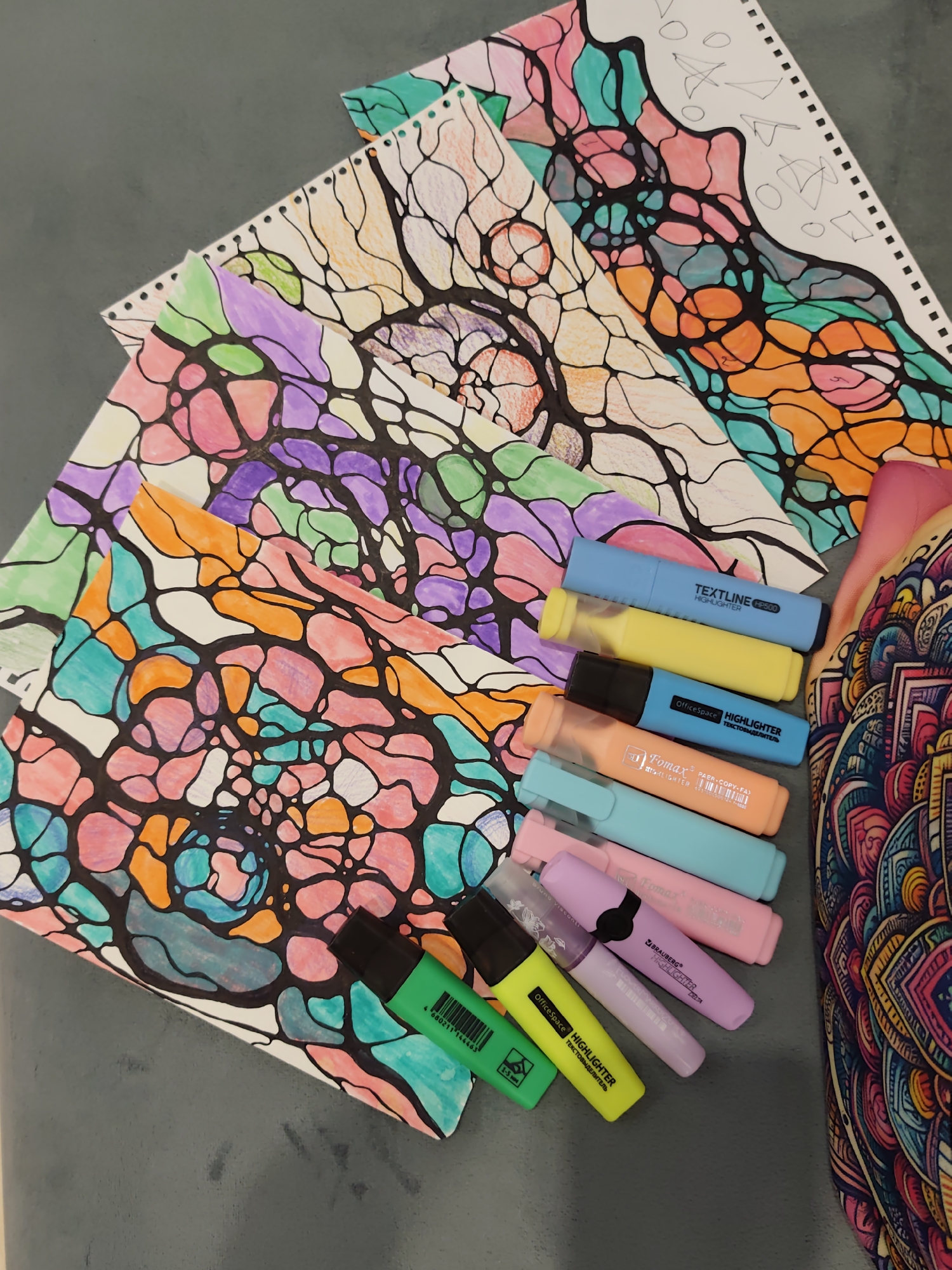 The image shows a close-up, overhead view of several sheets of paper with neurographic art, characterized by organic, interconnected lines and filled-in sections of various bright colors, including pink, orange, green, purple, and teal. Scattered on top of the papers are several highlighter pens in colors such as blue, yellow, orange, light blue, pink, and purple. The artwork and highlighters are arranged on a dark gray, textured surface. To the right of the artwork, a portion of a colorful, intricately patterned fabric, possibly part of a bag or clothing, is visible. The image does not contain any elements that can be used to determine its location in Sochi, Russia.