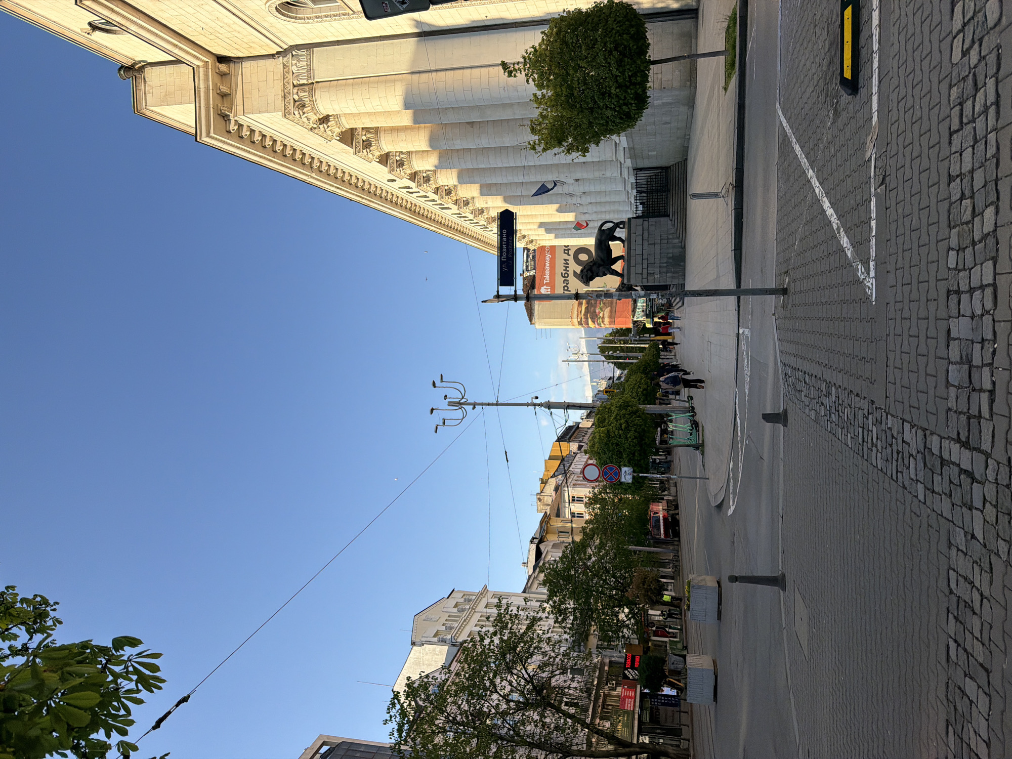 The image captures a street scene in Sofia, Bulgaria, taken from street level. The frame is vertically oriented, with the street running along the center, receding into the distance.
On the left side, a grand, light-colored building with classical architectural features, including columns and ornate detailing, dominates the view. Trees are positioned alongside it, partially obscuring some sections of the building.
A statue of a bronze lion stands near the building on the right. Adjacent to the lion and extending down the street, are commercial buildings adorned with signage, and various shops. Streetlights and power lines crisscross the sky above the street.
The street itself is paved with cobblestones. Pedestrians and vehicles populate the scene. The sky is clear, and the lighting suggests either early morning or late afternoon. The composition provides a balanced view of the city's architecture, street activity, and urban environment.