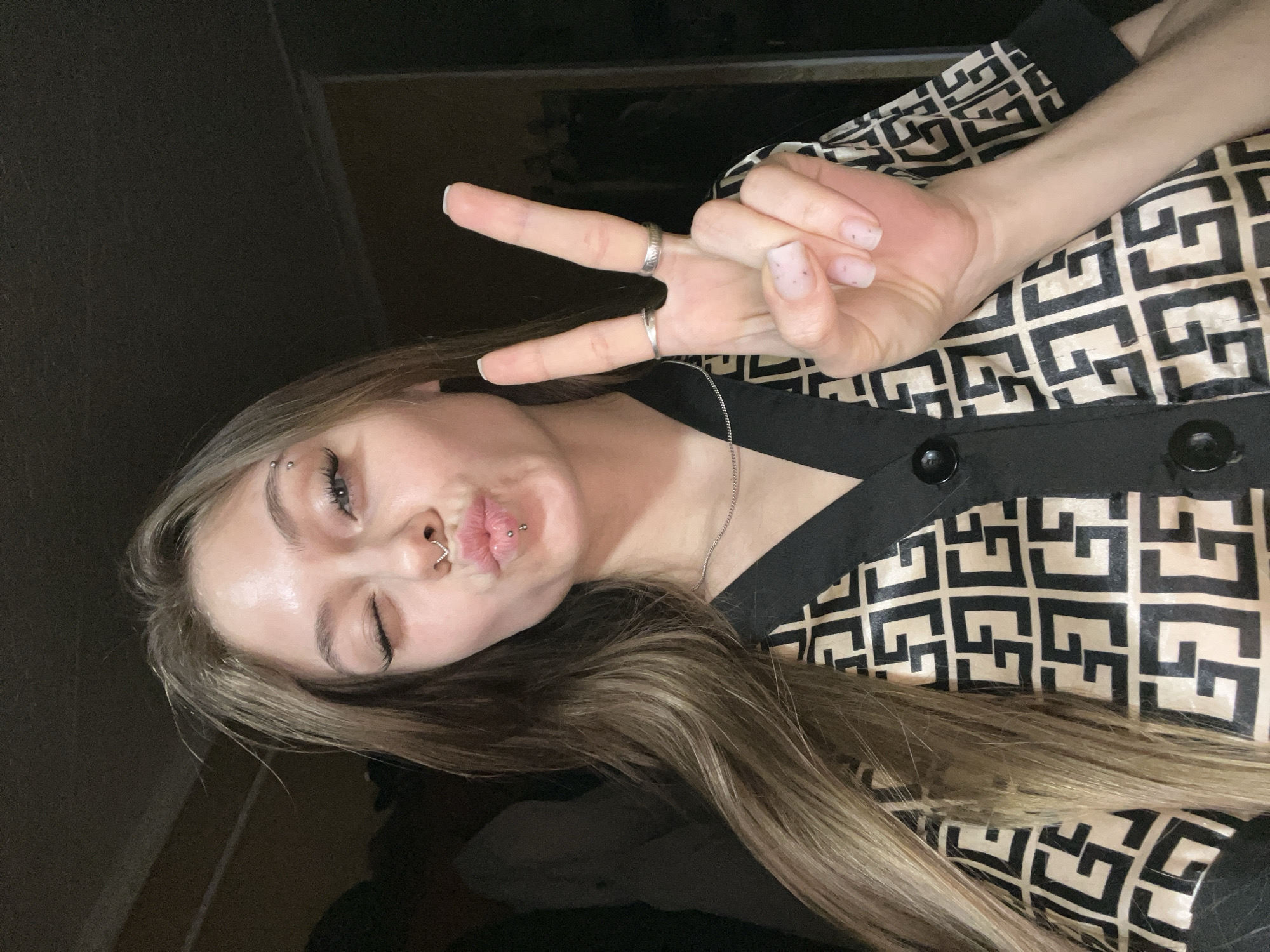 A young woman is posing indoors with her right eye closed and her lips pursed in a kiss. She is holding her left hand up, making a peace sign with her index and middle fingers. She has piercings in her eyebrow, nose, and lip. She is wearing a black and beige patterned shirt with black buttons. Her hair is long and blonde. The background is dark and indistinct. The image gives no indication of Фоки, Russia.