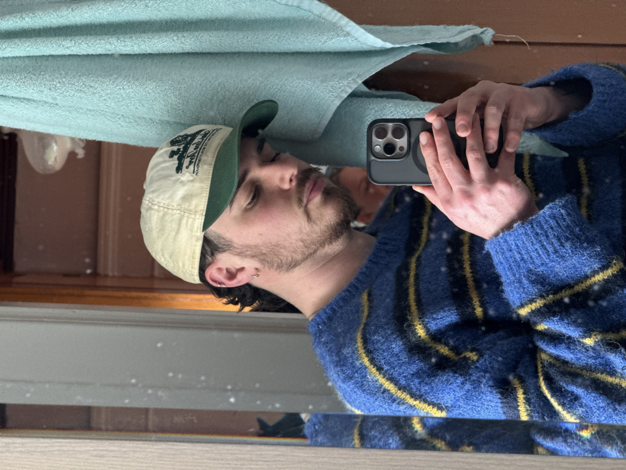 A man is taking a selfie in a mirror. He is wearing a blue sweater with yellow stripes and a baseball cap. A towel is hanging above him. The scene appears to be indoors, possibly in a bathroom. The city and specific location are unknown.