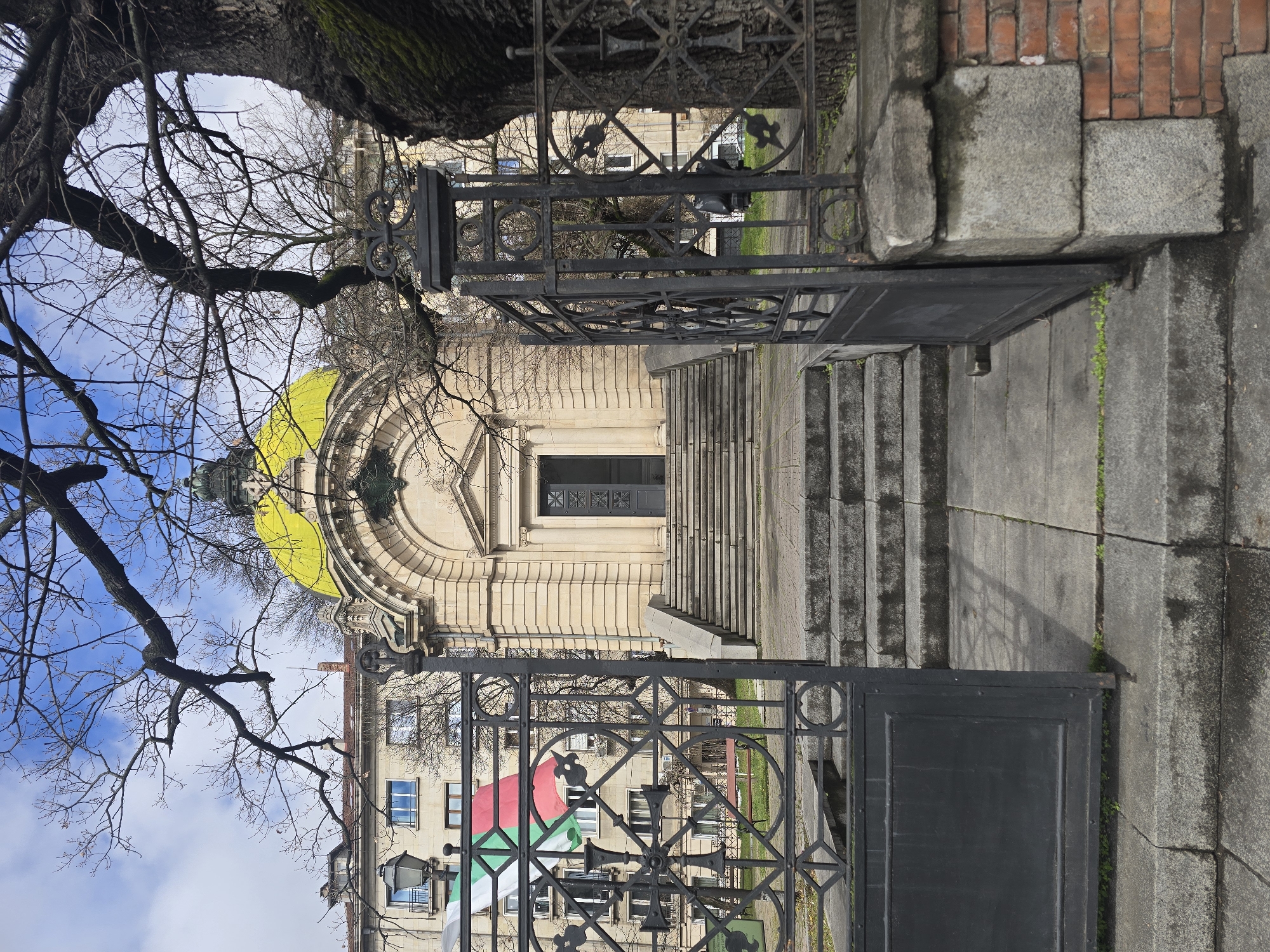 The image depicts a view through an ornate black iron gate toward a neoclassical building with a yellow dome in Sofia, Bulgaria. The building is accessed via a series of stone steps. Bare tree branches frame the scene at the top and left of the image. The sky is visible through the branches. The gate's design includes circular and diamond motifs. A building with a flag is seen through the gate. The pathway and surrounding stonework show signs of weathering and moss growth.