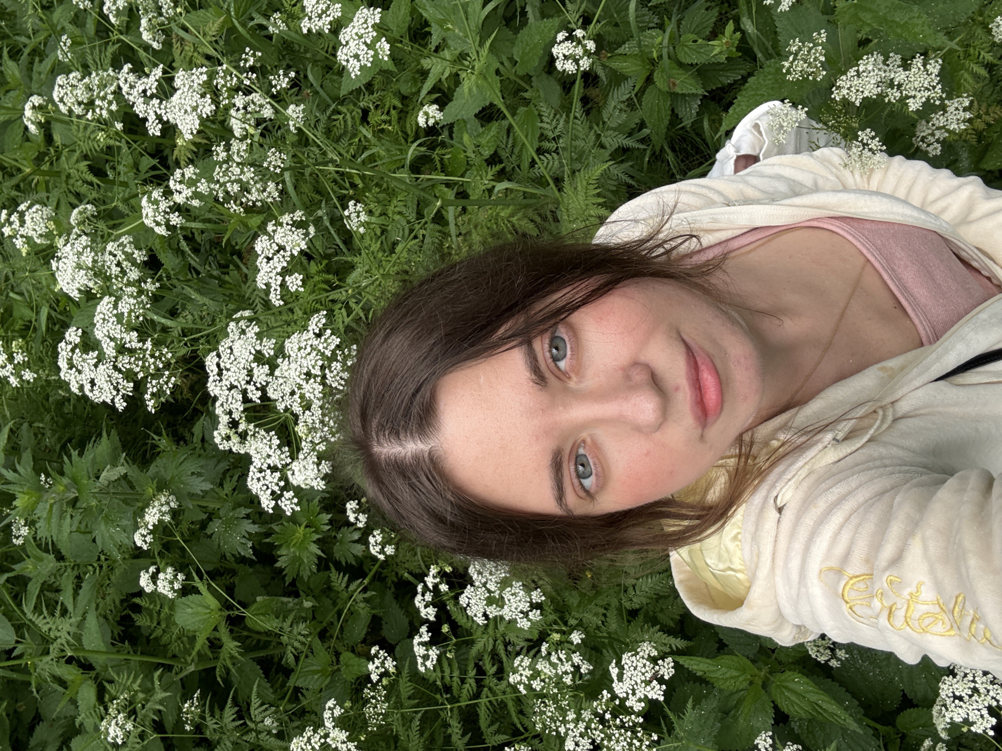 A young woman with brown hair and blue eyes is pictured outdoors in a field of green plants and white flowers. She is wearing a cream-colored hoodie over a pink tank top and a delicate gold necklace. The woman is looking directly at the camera. The background consists of lush green foliage interspersed with clusters of small white flowers. The image is a close-up, with the woman's face and upper body occupying the foreground, and the natural environment filling the rest of the frame.