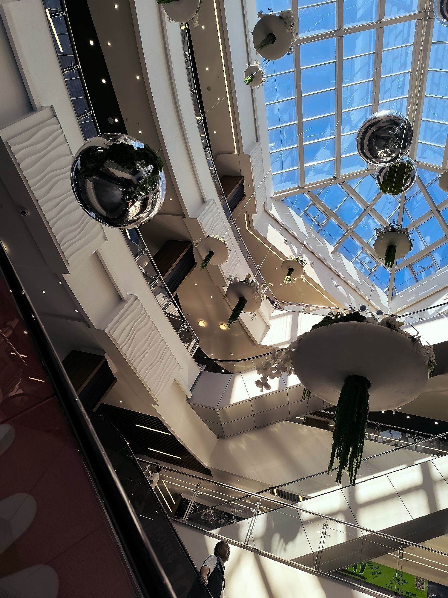 The image shows the interior of a modern shopping mall, likely in Qarağandı city administration, Kazakhstan. The perspective is looking upwards, revealing a multi-level structure with escalators and walkways. Large, reflective spherical ornaments and decorative hanging planters with greenery are suspended from the ceiling. A glass-paneled roof allows natural light to flood the space. In the foreground, an escalator is visible, with two individuals on it, one further up and the other closer to the camera. The architecture features clean lines, white walls, and glass railings. The overall ambiance is bright and spacious, characteristic of a contemporary retail environment.