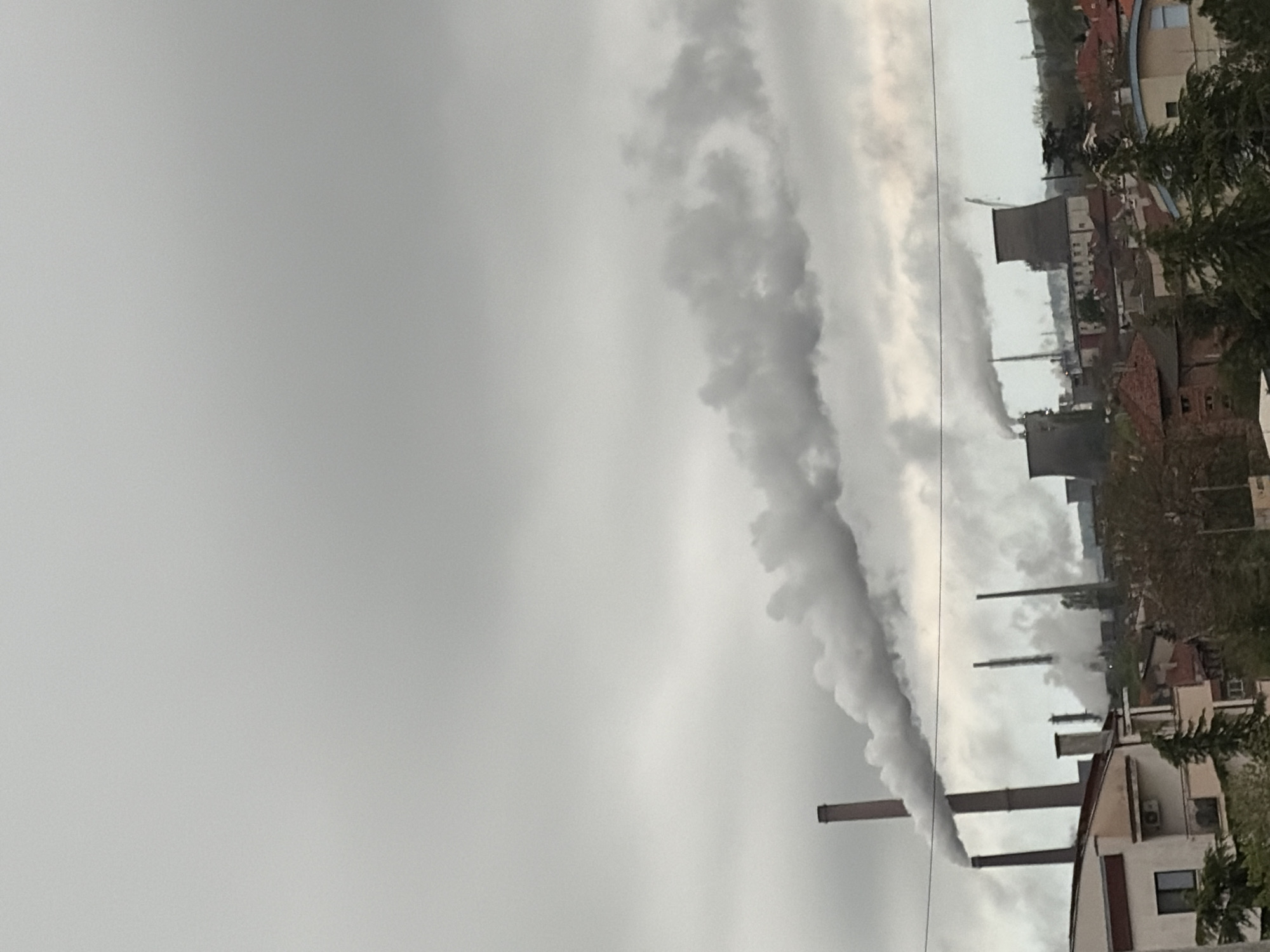 The image captures an expansive industrial landscape under a heavily overcast and grey sky. Numerous tall smokestacks and chimneys, varying in height and material (some brick, some dark grey), dominate the middle ground. From these structures, large plumes of thick, grey smoke or steam billow out, stretching horizontally across the scene and partially obscuring the sky.
Below the smoke and chimneys, a dense cluster of buildings forms what appears to be an industrial town or city. Many residential-style buildings with red or orange tiled roofs are visible, interspersed with larger, darker industrial structures, possibly cooling towers, particularly on the right side of the frame. In the immediate foreground on the bottom right, a white building with a brown roof and an external air conditioning unit is partially visible, alongside green foliage from trees and bushes. A thin black line, possibly a power line or cable, runs horizontally across the upper middle of the image, intersecting the smoke.
The scene depicts an active industrial area, with the continuous emission of smoke indicating ongoing operations. The weather is clearly cloudy and overcast, creating a muted, monochrome palette dominated by greys. The time of day is indeterminate due to the heavy cloud cover, but it is daylight. There are no visible people or discernible text in the image.