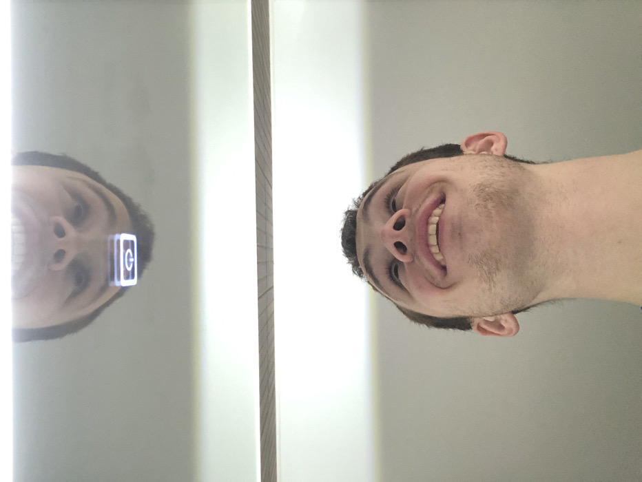 A man smiles, looking up at the camera. His head is slightly tilted to the side. To his left is a mirror, reflecting his face with a bright square over his forehead, showing a "G" logo. The background is mostly off-white, and a vertical wooden element separates the man from his reflection.