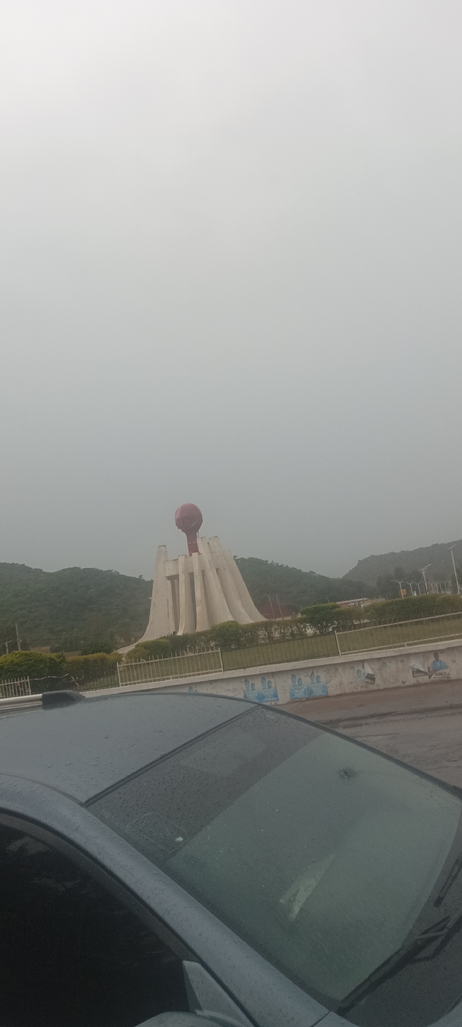 The image depicts the National War Memorial in Abuja, Nigeria, partially obscured by a car's windshield in the foreground. The memorial consists of a cluster of white columns supporting a red spherical structure. Green hills are visible in the background. The sky is overcast, suggesting rain or heavy cloud cover. A concrete wall with a metal fence sits in front of the memorial, and indistinct images appear to be painted or displayed on the wall. The view is captured from inside a vehicle, evident by the car's hood and windshield with rain droplets.