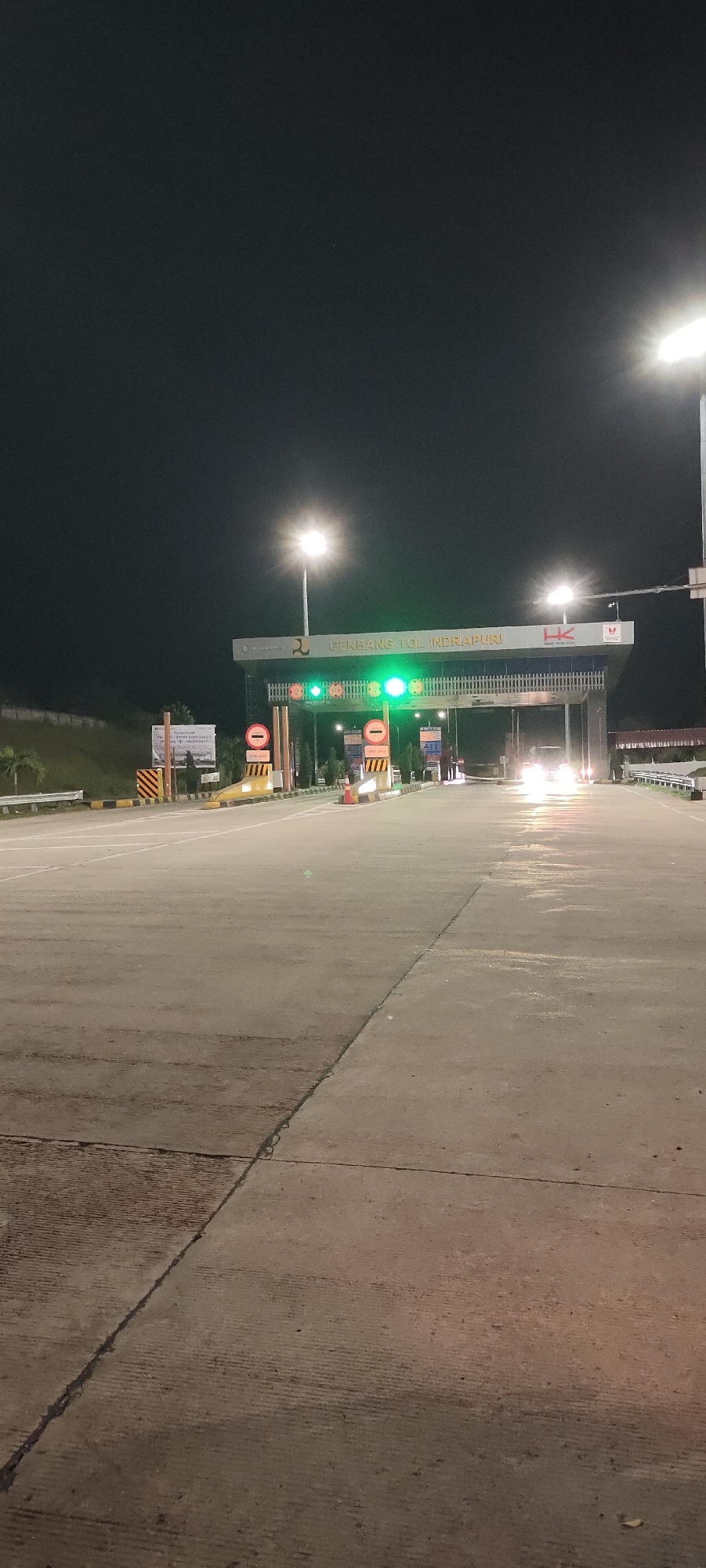 The image captures a nighttime view of the Indrapuri Toll Gate in Sihomlhok, Indonesia. The scene is illuminated by overhead streetlights, casting a bright glow on the wide concrete road and the toll plaza structures. The toll gate itself is a prominent feature, with a large overhead sign displaying "GERBANG TOL INDRAPURI" and the "HK" logo. Green traffic lights are visible, indicating that the toll lanes are open. Several vehicles are present, with headlights visible as they approach or pass through the toll booths. The ground is wet, reflecting the artificial light and suggesting recent rain. The surrounding environment is dark, with a hint of foliage on the left side of the image and a guarded embankment. The overall atmosphere is that of a functional, well-lit, and active toll collection point during nighttime hours.