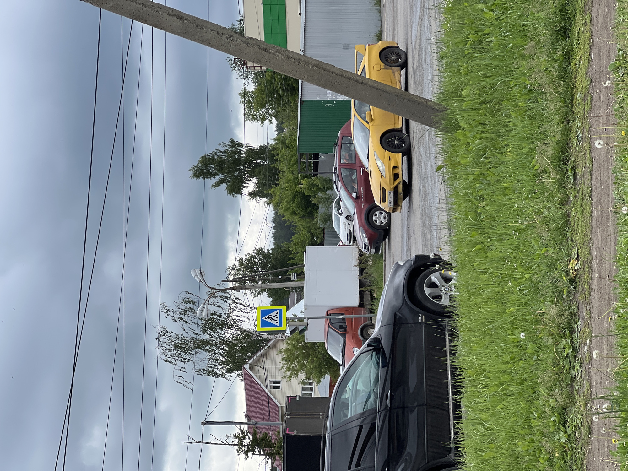This image depicts an outdoor scene in a mixed-use area of Izhevsk, Udmurt Republic, Russia, identified by the "18" region code on a vehicle license plate. The sky is completely overcast.
In the foreground, on the left, a patch of green grass with dandelions and a dirt path is visible. The front passenger side of a black BMW X5 SUV is partially visible, positioned over the grass. On the far right, a concrete utility pole angles diagonally upwards, partially obscuring the view.
The midground features a paved area or road. A yellow Toyota Celica, modified with a large rear spoiler and black wheels, is positioned centrally, facing right. Its license plate reads "C380PX 18". Behind the Celica, a red Dacia/Renault Sandero Stepway crossover with roof rails is visible, also facing right. Further left and behind the black BMW, an orange-cab white-box truck, resembling a GAZelle, is parked. To its left, a dark truck with an open rear container is partially visible. A yellow and blue square pedestrian crossing sign is mounted on a pole near the center-left, adjacent to a street light. A white sedan is partially visible behind the red crossover on the far left.
The background presents a varied environment. A two-story house with a red roof and light-colored siding is visible behind the orange-cab truck and pedestrian sign. To the right of the house, a series of commercial or industrial buildings with green facades and white siding are present. Scattered throughout the background are numerous deciduous trees with green foliage. Multiple black overhead power lines crisscross the entire upper portion of the image against the gray sky. No human figures are clearly visible.