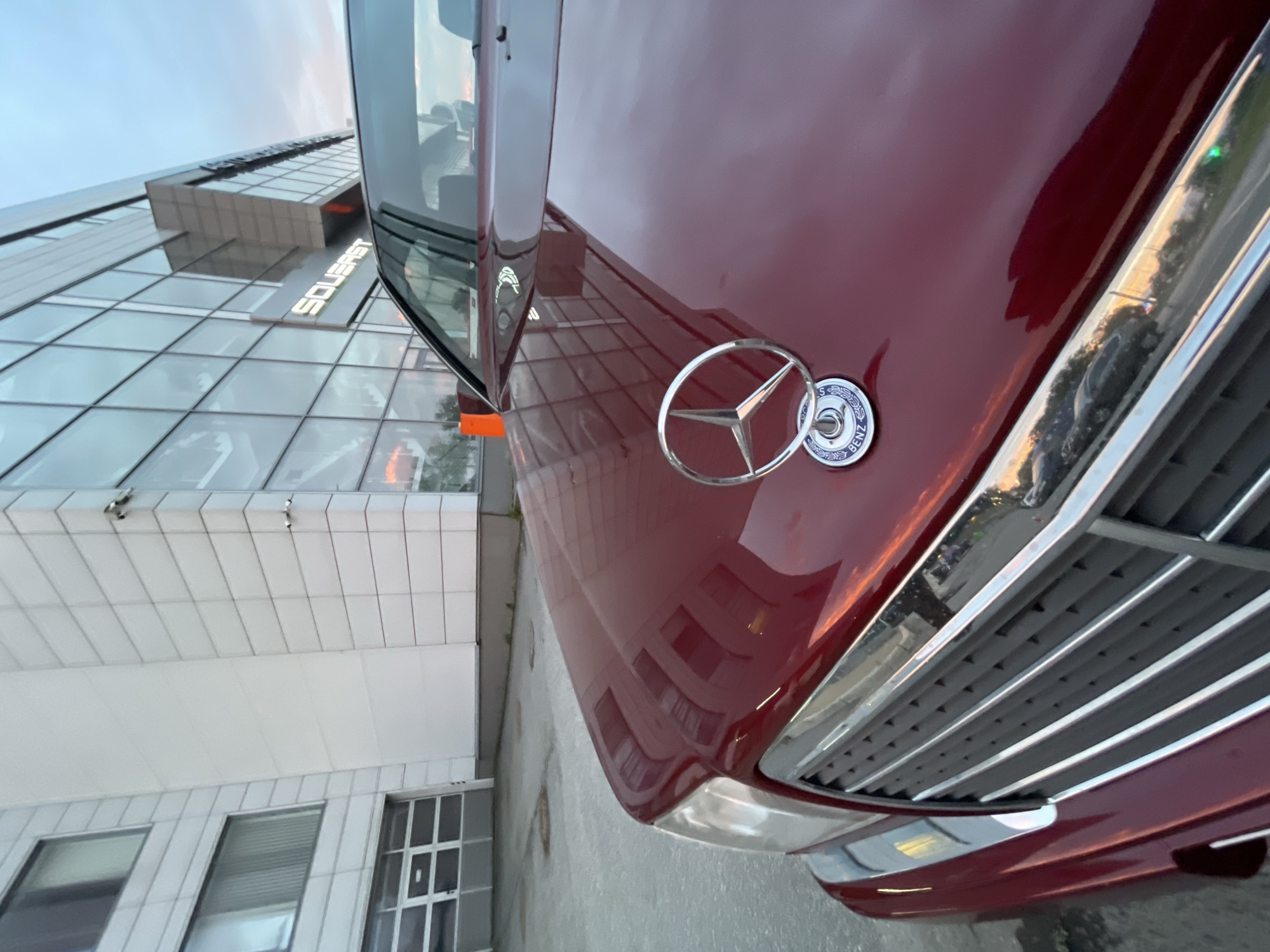 The image shows the hood and front grille of a dark red Mercedes-Benz car parked outside a modern building with a glass facade. The building has the word "SOUERAST" illuminated in white letters. The sky is overcast, and there are reflections of the sky and the building on the car's polished surface. The car's emblem, a three-pointed star within a circle, is prominently displayed on the hood. The environment appears to be an urban setting, possibly a commercial or office area.