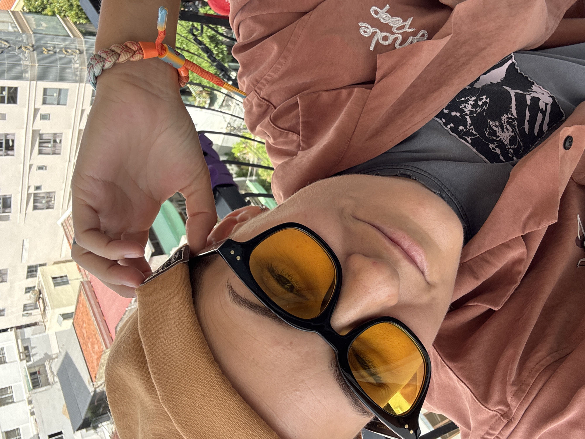 In a close-up, selfie-style shot taken on a balcony, a person is partially visible, wearing a brown beret, black-framed yellow-tinted sunglasses, and a rust-colored button-up shirt over a dark gray t-shirt. A colorful braided bracelet adorns their wrist. The background reveals a view of urban rooftops with various buildings, suggesting a city setting, likely Nha Trang Ward, Vietnam, given the architectural style and foliage. The person's left hand is raised, touching the brim of their beret. The lighting indicates it is daytime.