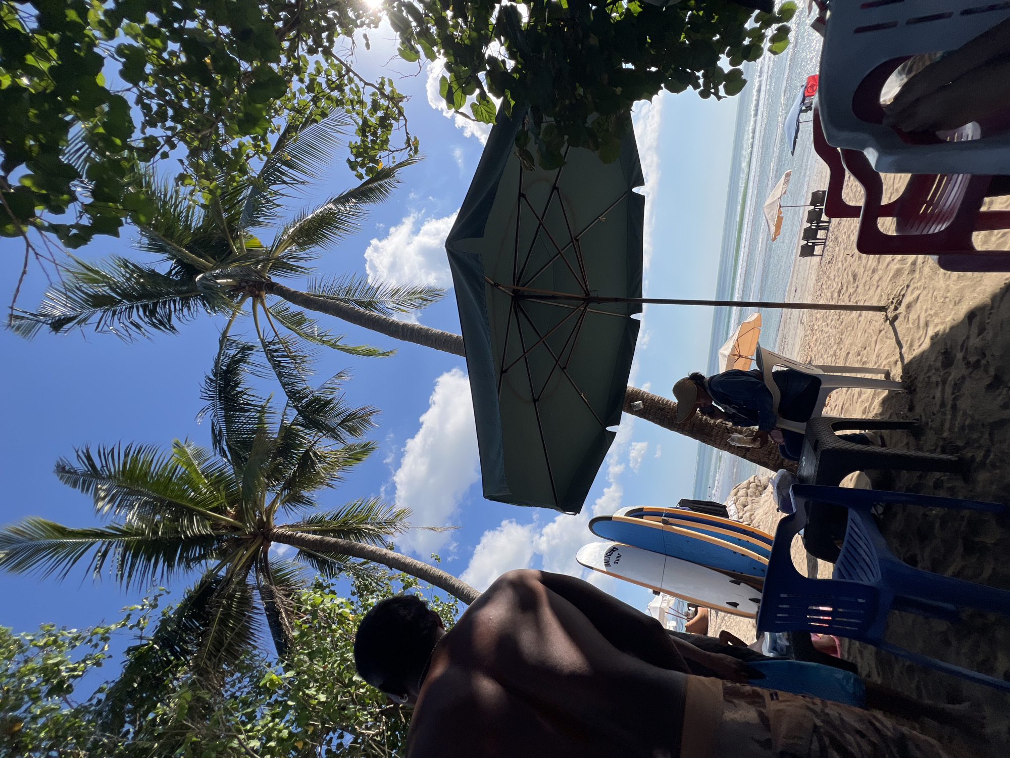The image depicts a beach scene in Kuta, Indonesia, under a clear blue sky with scattered clouds. In the foreground, the back of a person is visible, facing away from the camera and towards the ocean. To the left, several surfboards are propped up on the sand. In the middle ground, a large green umbrella shades a table and chairs, where a person wearing a hat and denim jacket is seated, holding what appears to be food. Further down the beach, more umbrellas and chairs are set up. Palm trees frame the top of the image, and foliage from trees on the right creates a dappled shadow effect, with sunlight glinting through the leaves. The ocean stretches to the horizon, with gentle waves rolling onto the shore. The overall atmosphere suggests a relaxed beachside setting.