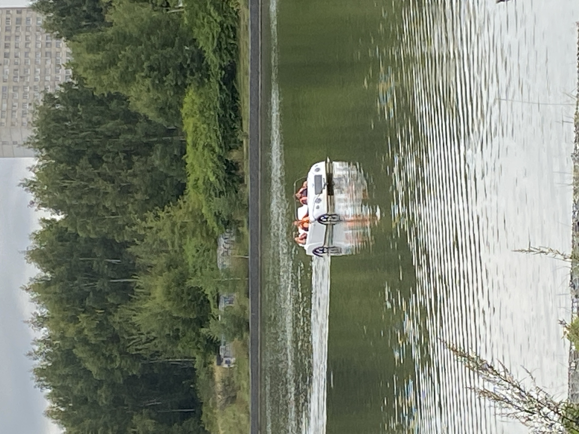 An aerial view shows a white pedal boat with two people wearing orange life vests on a body of water. The boat is shaped like a small convertible car. The water has ripples and reflections, with some plant life visible along the shore. In the background, there is a dense line of trees, and beyond that, a tall, light-colored building with many windows. The sky is overcast. The location is Unknown, Unknown.