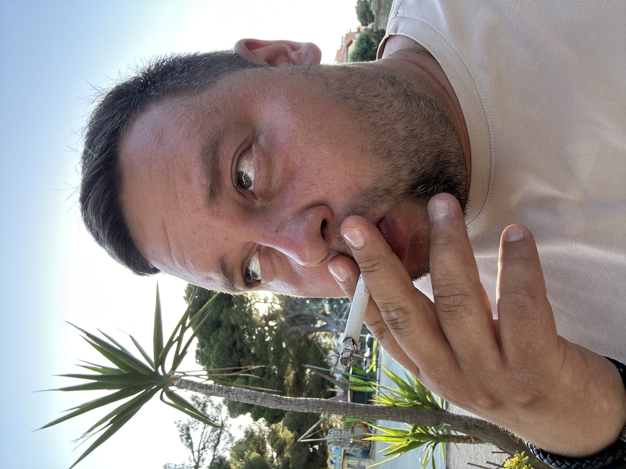 A man is smoking a cigarette outdoors, in what appears to be a park or recreational area with palm trees and other greenery. He is looking up and to his left, with his right hand holding the cigarette to his mouth. The background shows trees, what looks like playground equipment, and a clear sky. The lighting suggests it is late afternoon or early evening. The image is a close-up, focusing on the man's face and hand. The location is Sant Feliu De Guíxols, Spain.