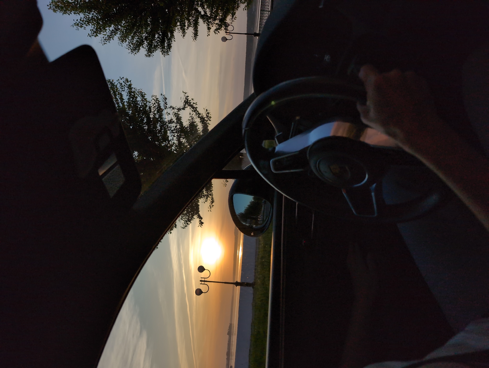 A person is driving a car in Votkinsk, Russia, as indicated by the presence of a lamppost typical of the city's waterfront. The photograph is taken from inside the vehicle, looking out towards the sunset over a body of water, presumably the Votkinsk Reservoir. The steering wheel and the driver's hand are visible, suggesting that the driver is actively navigating the vehicle. Tree foliage is seen above the car's roof, indicating the presence of greenery nearby. The car's side mirror reflects the sunset and the shoreline, further contextualizing the location.