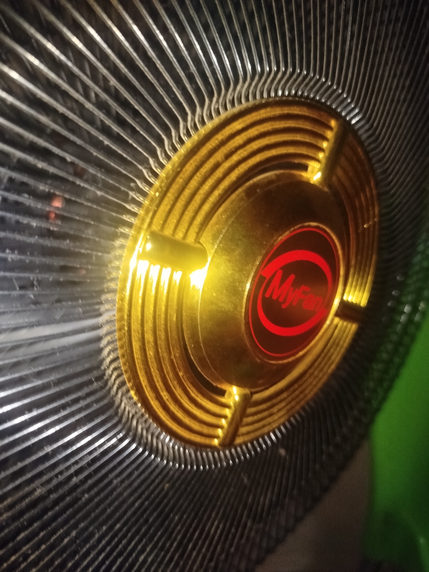 This is a close-up, high-angle shot of a CPU cooler fan. The fan has a gold-colored metal heatsink with numerous fins radiating outwards. In the center of the heatsink is a circular emblem with a red border and a red logo inside. The text "MyFan" is visible within the emblem, written in a stylized red font. The fan is positioned against a dark background, with some bright, out-of-focus green in the upper right corner, suggesting it's part of a larger electronic device. The lighting is harsh and direct, creating strong reflections and highlights on the metal surfaces, particularly the gold central part. Some dust or debris is visible on the fins of the heatsink. There is no visible activity or event taking place; the image focuses solely on the static object. The overall impression is one of mechanical detail and the metallic sheen of the components. The text "MyFan" indicates the brand or model of the fan.