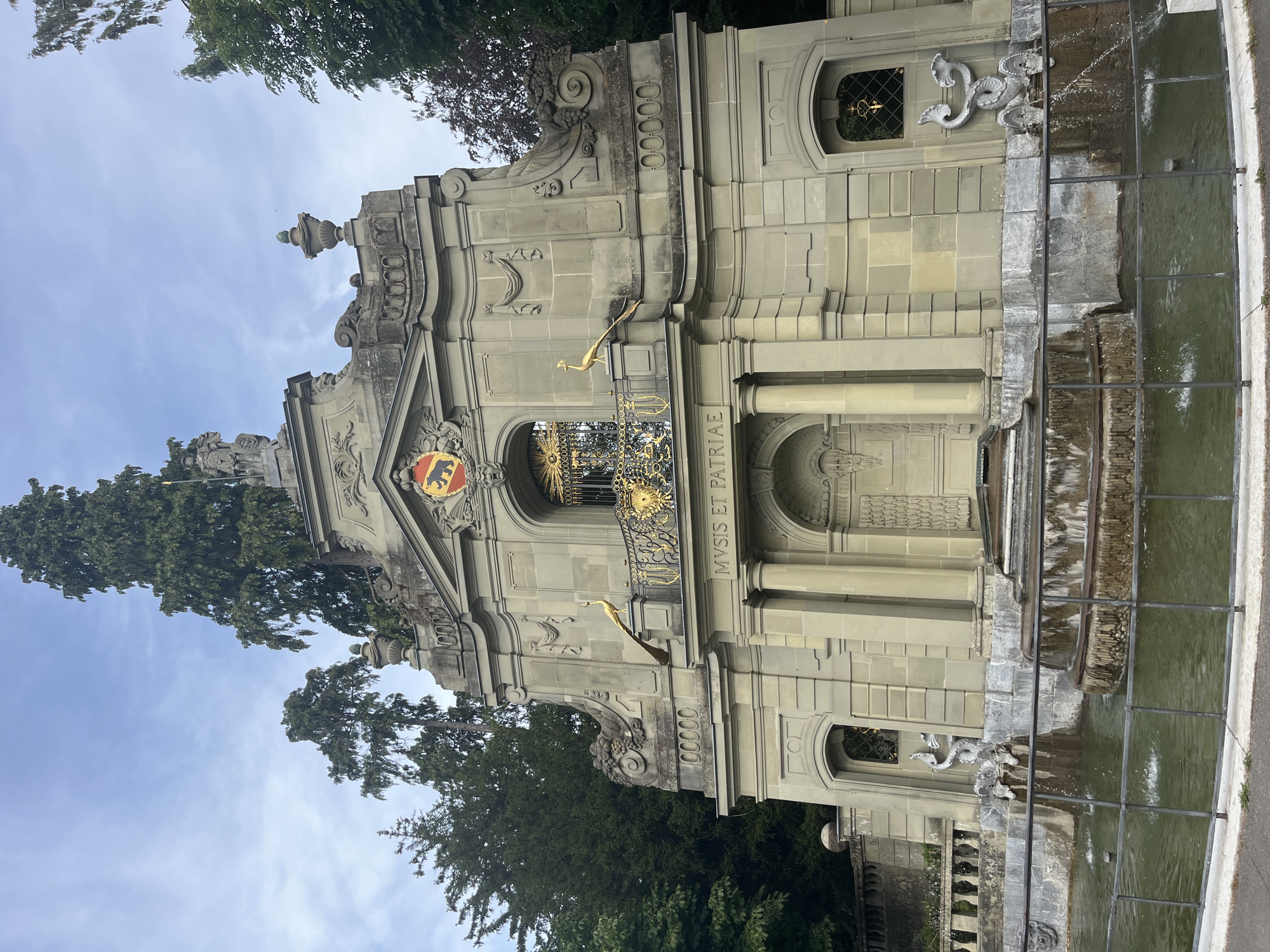The image shows the Zytglogge fountain in Bern, Switzerland. The fountain is a Baroque-style structure made of sandstone, featuring elaborate carvings and sculptures. In the center of the fountain is a tall, tiered column topped with a statue of a figure. Flanking the column are two golden peacocks perched on a balcony. Above the balcony, a crest with a bear is displayed. The inscription "MVSIS ET PATRIAE" is visible on a stone plaque. The fountain itself has multiple levels of water cascading down into a pool. On either side of the fountain's base, there are two sculpted sea creatures from which water flows. The fountain is surrounded by a metal fence. In the background, tall evergreen trees and a cloudy sky are visible.