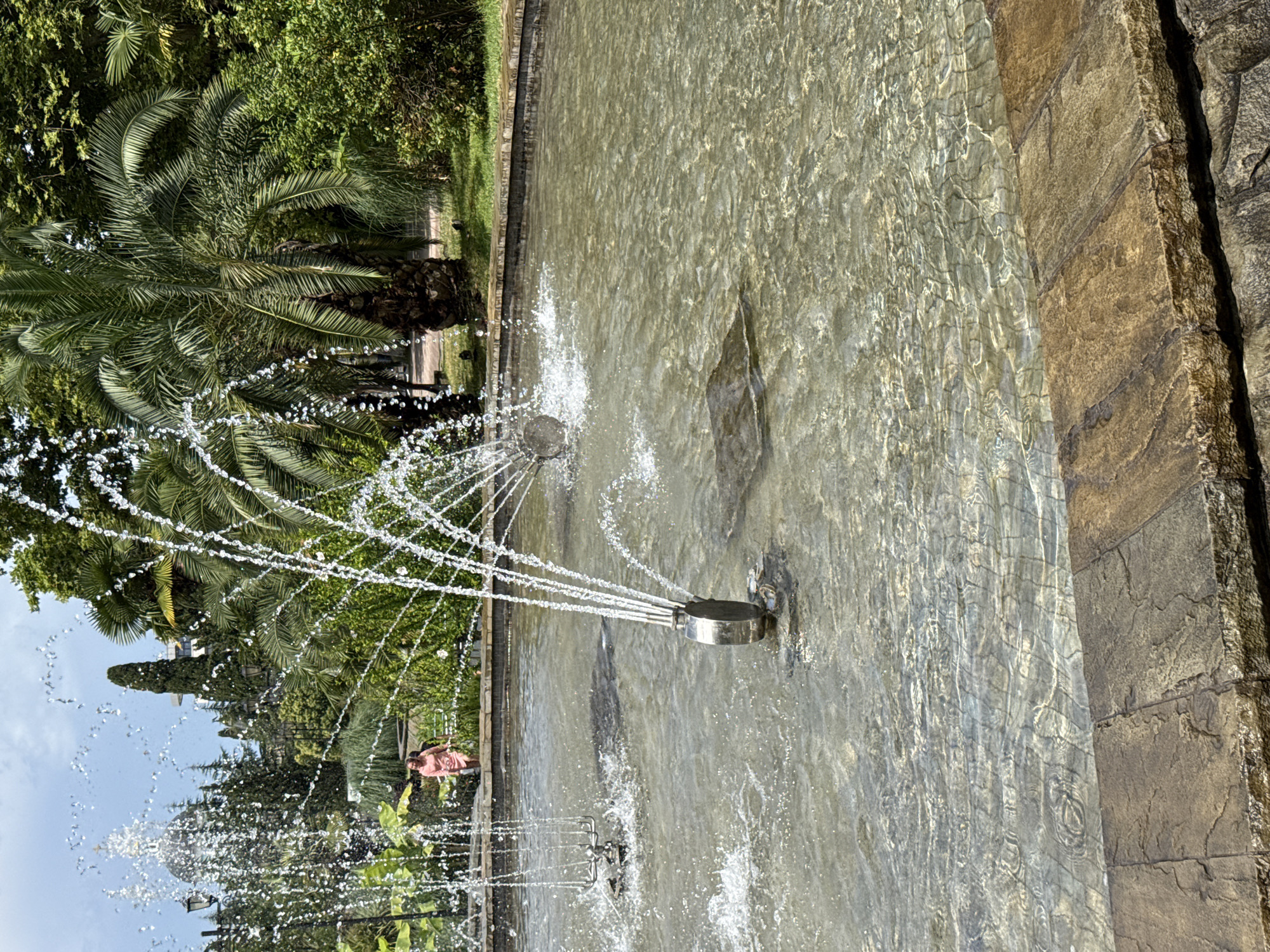 In Sochi, Russia, a round fountain with multiple water jets is the central focus of this image. The water is clear and shallow, with visible tiling beneath the surface. The fountain's metallic spray heads are modern and artistic, with one featuring multiple slender spouts and another with a more branched design. In the background, a woman in a pink dress walks along a stone pathway. Lush green foliage, including palm trees and other tropical plants, creates a vibrant, natural backdrop. The scene suggests a public park or garden setting.