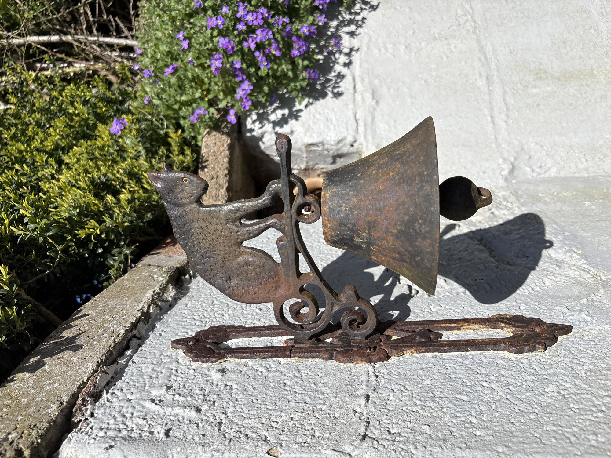 The image shows a rusty, cast iron cat-themed door bell. The cat figure is attached to a decorative bracket holding a weathered, cone-shaped bell with a spherical clapper. The bell assembly is mounted on a whitewashed brick wall, likely a fence or the side of a building. Adjacent to the bell are green bushes and purple flowers. Given the prompt, this scene is presumed to be located in Snailbeach, United Kingdom.