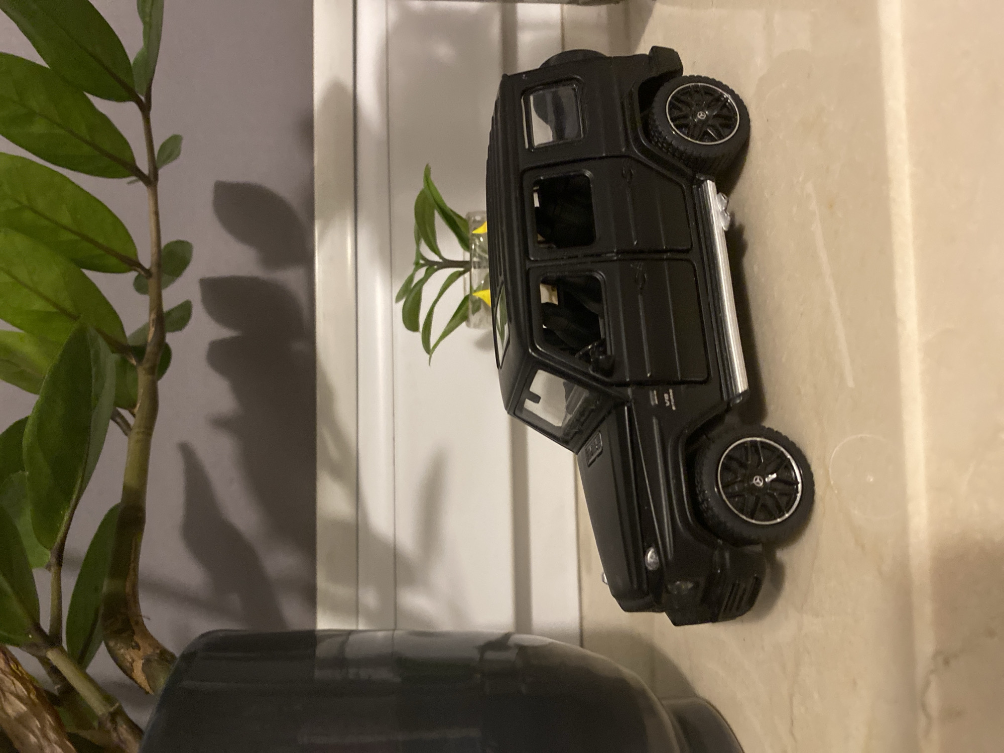 A black toy Mercedes-Benz G-Class is positioned on a marble-patterned surface. The toy car faces right, with its rear wheel visible, featuring a black rim with silver accents and a Mercedes-Benz logo. To the left of the car, a dark gray planter holds a green leafy plant, casting a shadow on a gray wall behind it. A smaller green plant in a glass vase is visible in the background, to the right of the toy car. The environment appears to be indoors, on a shelf or table. There is no indication of the city of Łódź, Poland, or any specific urban location.