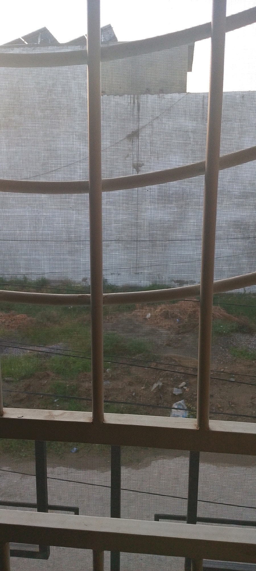 The image, taken from behind a barred window with a mesh screen, looks out onto a construction site or undeveloped land. In the foreground, vertical metal bars and horizontal bars of a light brown or beige color are prominent. The mesh screen is a fine grid pattern that covers the entire view.
Beyond the bars and screen, the scene consists of uneven ground with patches of green grass and areas of exposed brown soil, suggesting construction activity or neglect. There are piles of dirt or debris. Several dark horizontal lines, possibly electrical wires or cables, are visible crisscrossing the middle ground. In the far background, a large, light-colored wall or structure is visible, with some indistinct shapes on top that might be roofs or structures. The sky above appears overcast or very bright, as there is a strong wash of light.
The image does not contain any people or discernable actions. The environment appears to be outdoors, in a location that is undergoing some form of development or has been left undeveloped. Based on the provided metadata, the location is Attock, Pakistan.