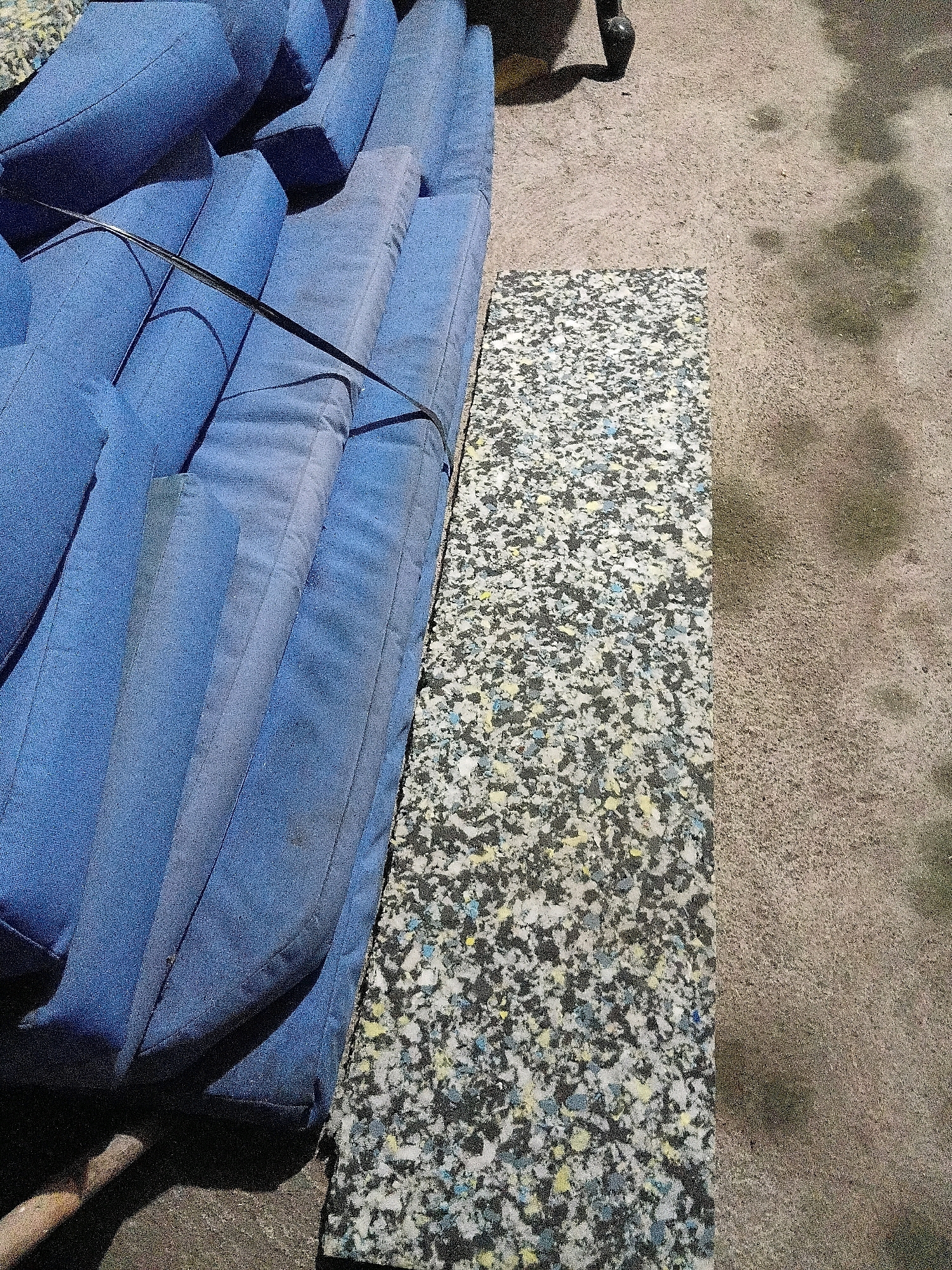 A pile of blue upholstered cushions is stacked on top of each other, tied together with a black strap. Beside the cushions lies a rectangular piece of foam padding. The scene appears to be outdoors on a dusty, textured ground, possibly a sidewalk or vacant lot. Shadows suggest it is daytime. The ground is a light brown color and has some dark smudges. There is a shadow of an object, possibly a chair or table leg, on the ground to the upper right of the cushions. The location is identified as Cikal, Indonesia.
