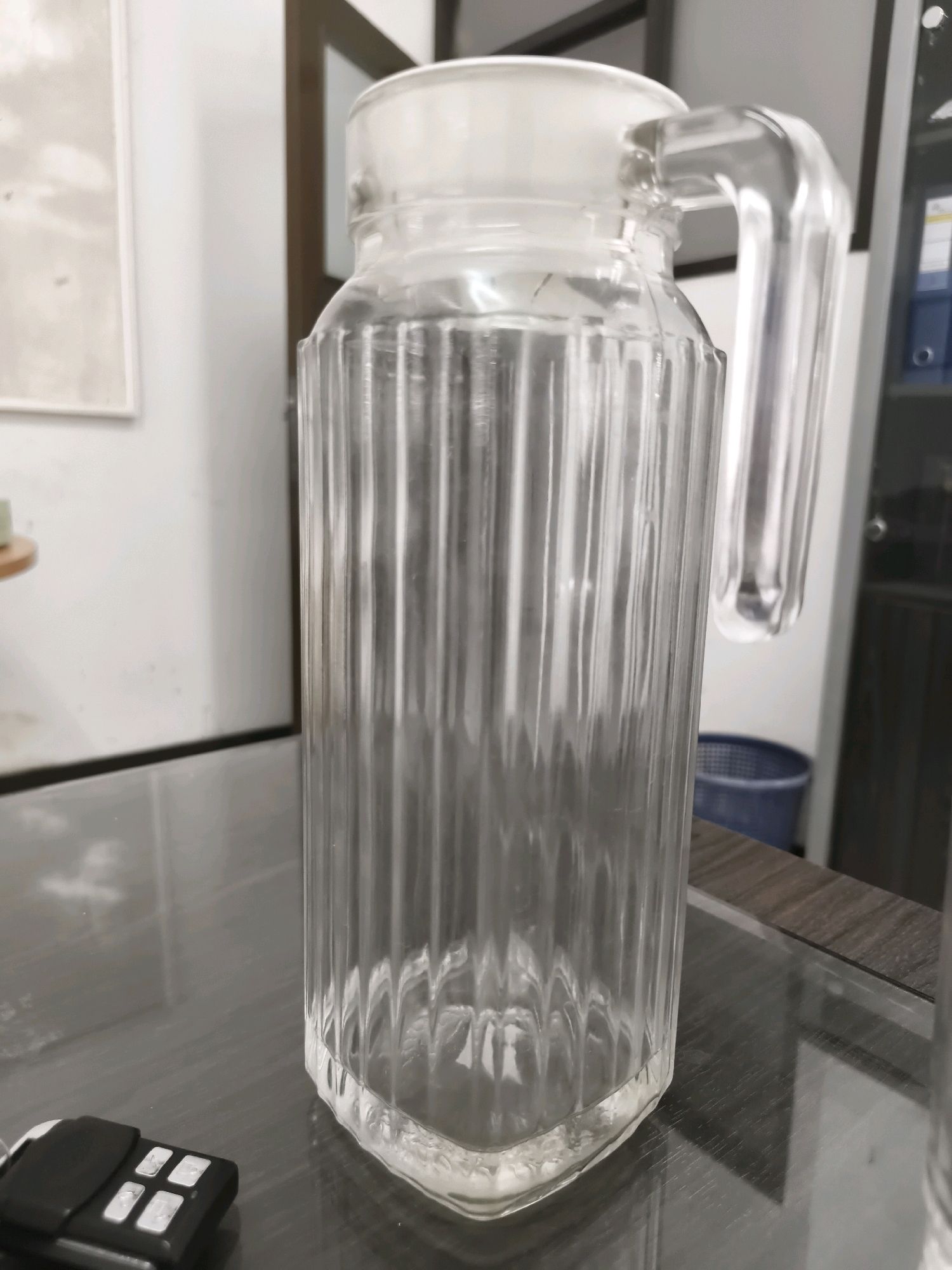 A clear glass pitcher with vertical ridges and a clear glass handle sits on a reflective surface. The pitcher is tall and cylindrical with a faceted base. A white lid is visible at the top of the pitcher. In the foreground to the left, a black key fob with several white buttons rests on the surface. In the background, a framed picture hangs on a white wall, and a blue plastic bin is visible. The scene appears to be indoors, possibly in a home or office setting. The location is not specified in the image.