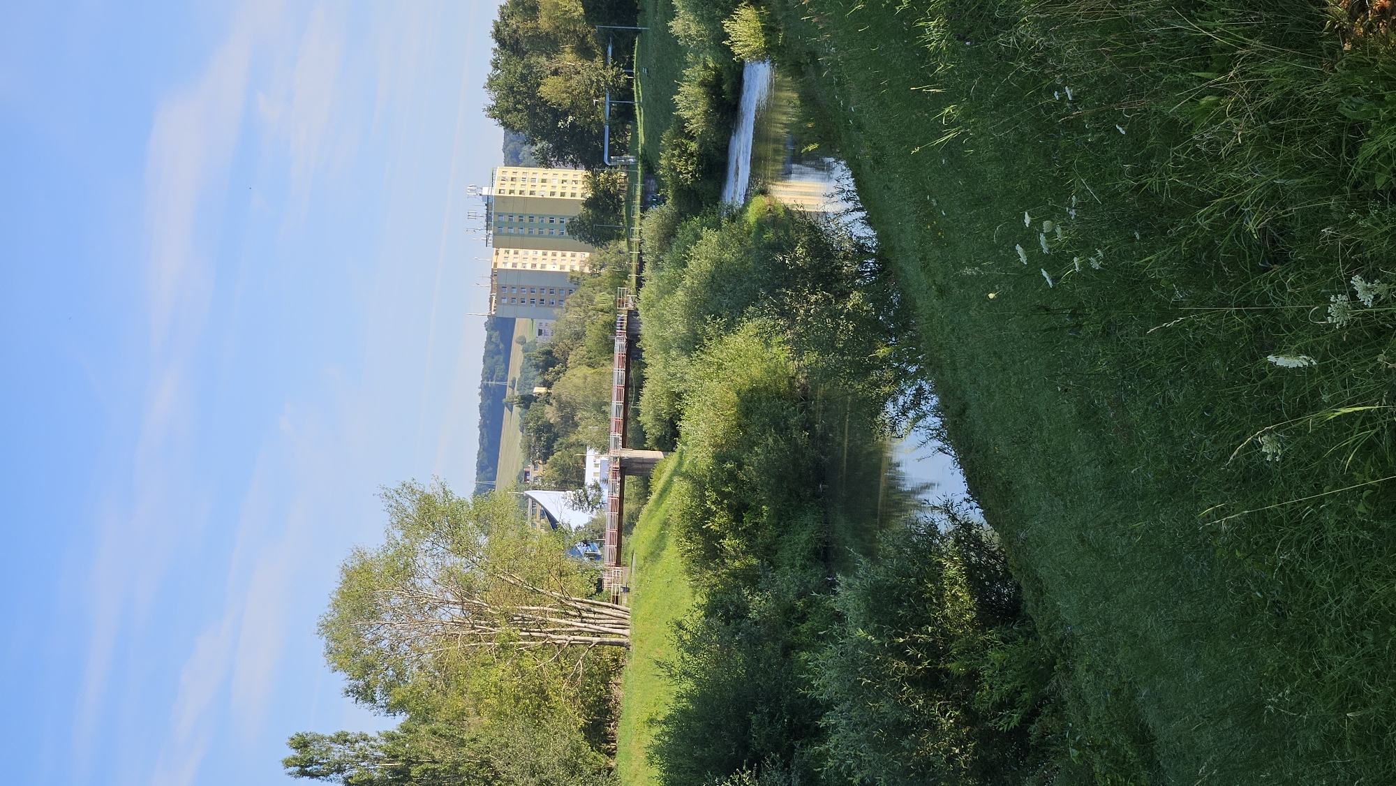 The image captures a serene riverside scene with tall buildings in the background, located in the city of Prešov, Slovakia. In the foreground, a grassy slope leads down to a winding river, its surface reflecting the clear blue sky and surrounding greenery. Lush bushes and trees line the riverbanks, creating a vibrant natural border. A pedestrian bridge with red railings spans the river in the middle ground, connecting the green banks. Beyond the bridge, two modern apartment buildings, one yellow and the other green, rise prominently against the horizon. The architecture of these buildings suggests a residential area. The overall environment is peaceful, with no people or animals visible in the frame. The lighting suggests it is either early morning or late afternoon, with the sun casting a gentle glow on the landscape. The distant hills, covered in trees and some patches of grass, add depth to the scene.