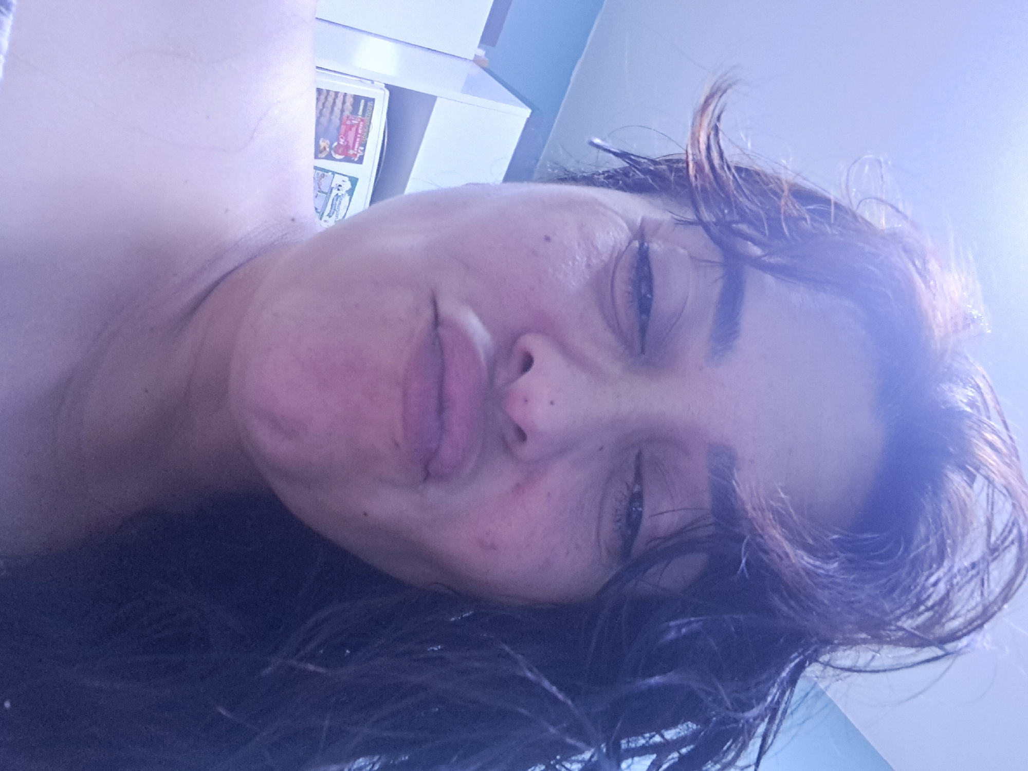 The media file is a close-up, tilted selfie of a person. The subject's face is the primary focus, with their eyes partially closed and their lips pursed. Their dark hair is wet and messy, framing their face and falling across their shoulder. The lighting is bright and appears to be from overhead, casting a cool, purple-blue hue over the scene, suggesting an indoor setting with artificial or bright natural light. In the background, to the left, parts of white shelving units are visible, with some items on them, including what appears to be a stack of magazines or cards with colorful imagery. The overall impression is of a casual, personal moment captured at close range. The visible text, if any, is not legible due to the image quality and angle. The context suggests a location in Manisa, Turkey, though no specific visual cues confirm this within the image itself. The person's expression is somewhat ambiguous, perhaps conveying relaxation or playfulness. The time of day is not ascertainable, but the lighting suggests it's not the dark of night.