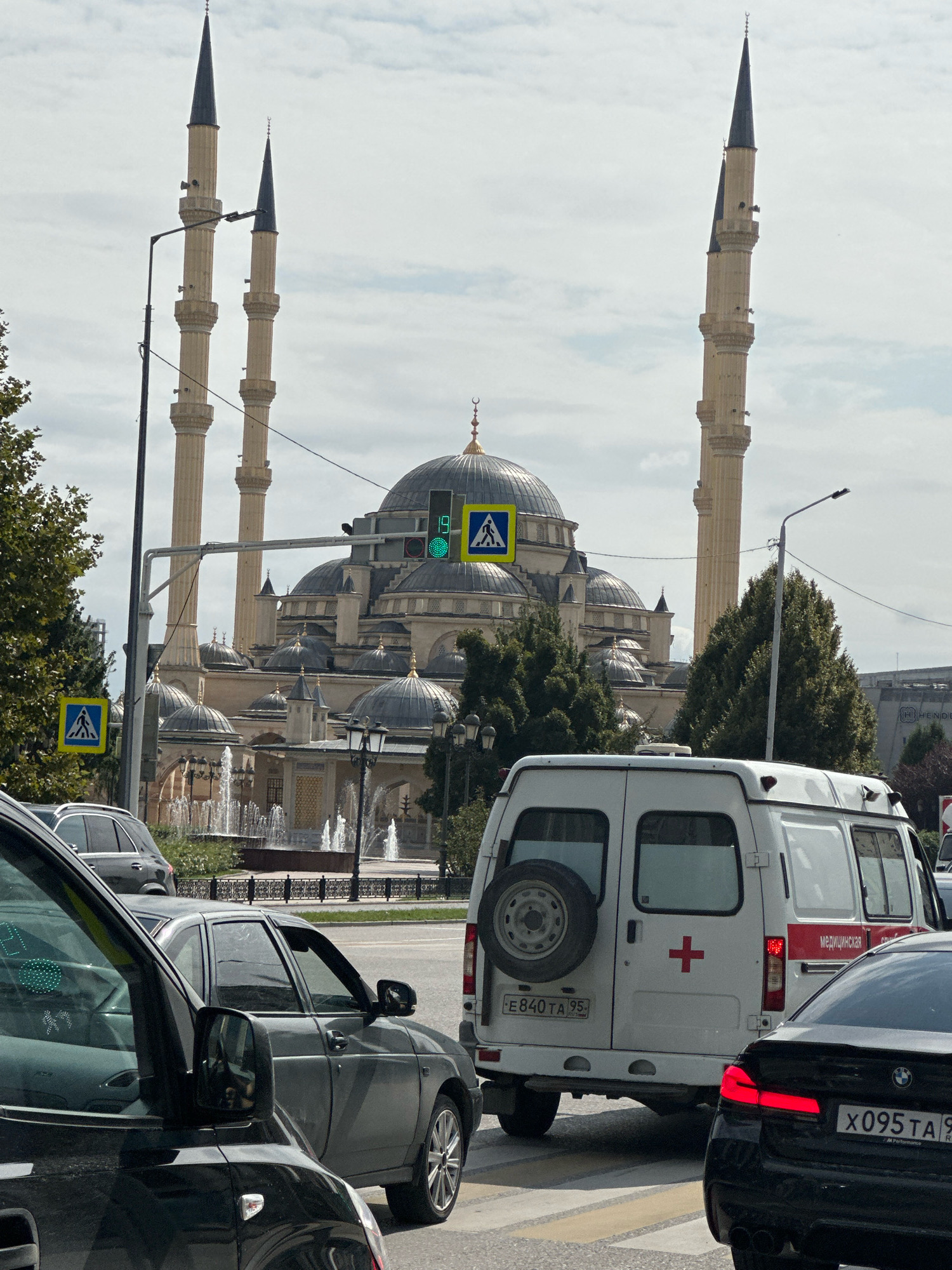 The image shows a street scene in Grozny, Russia, with the Akhmad Kadyrov Mosque prominently in the background. The mosque features multiple domes and tall minarets. In the foreground, traffic is backed up at an intersection. A white ambulance with a red cross and the word "МЕДИЦИНСКАЯ" (medical) is visible, alongside other vehicles including a black BMW. A pedestrian crossing is marked on the road. Traffic lights are green. Trees and streetlights line the road. The sky is overcast with clouds.