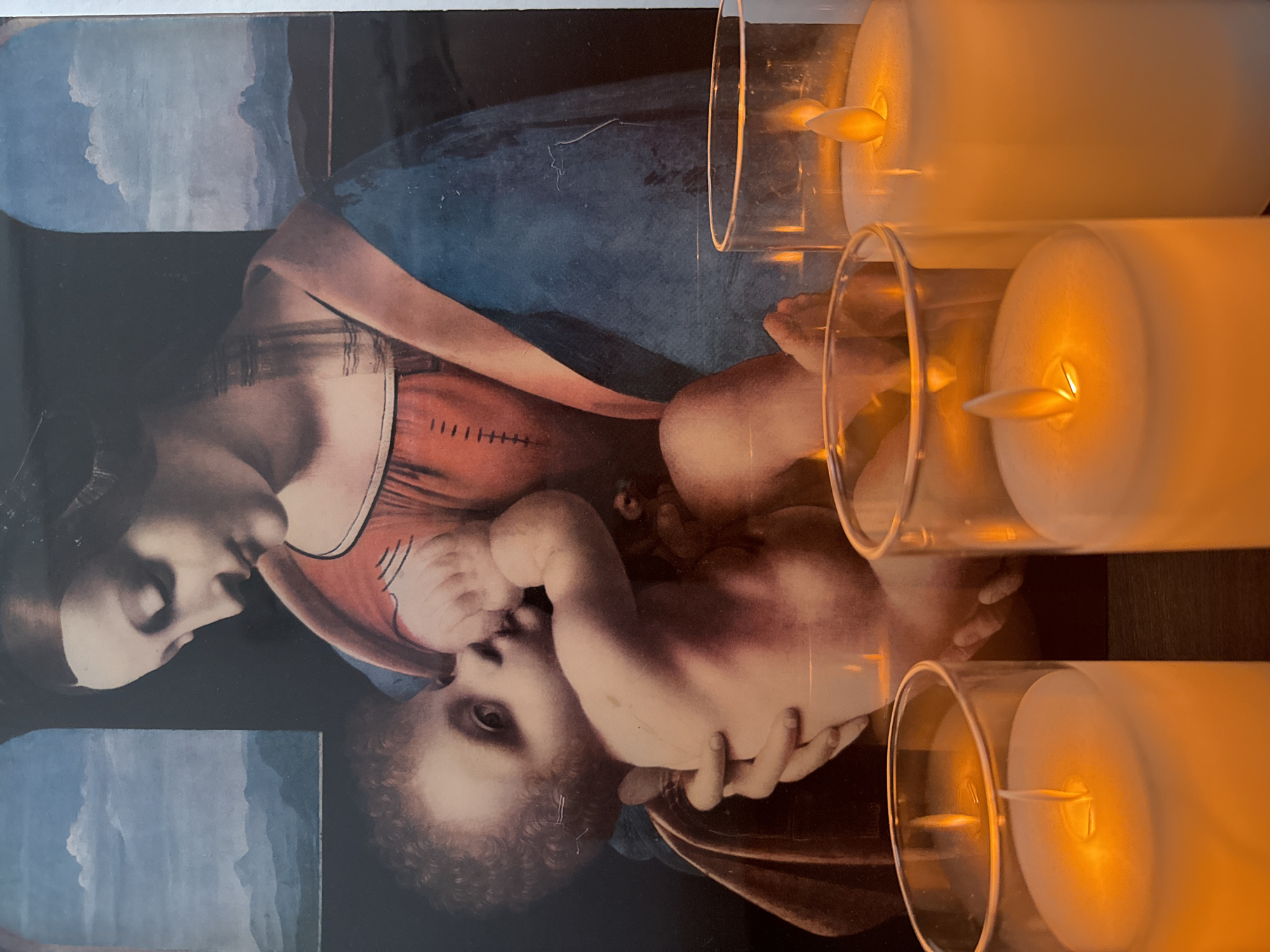 A print of a Renaissance painting depicting the Virgin Mary and the infant Jesus rests next to three lit electric candles in glass holders. The painting is a cropped view of a broader scene, showing Mary with dark hair and pale skin, cradling Jesus in her arms. He is a fair-skinned baby with curled hair. The background includes a dark blue sky and a landscape. The three cylindrical candles are arranged vertically, with the top one being the clearest. The environment is dimly lit. Votkinsk is not discernible from the image.