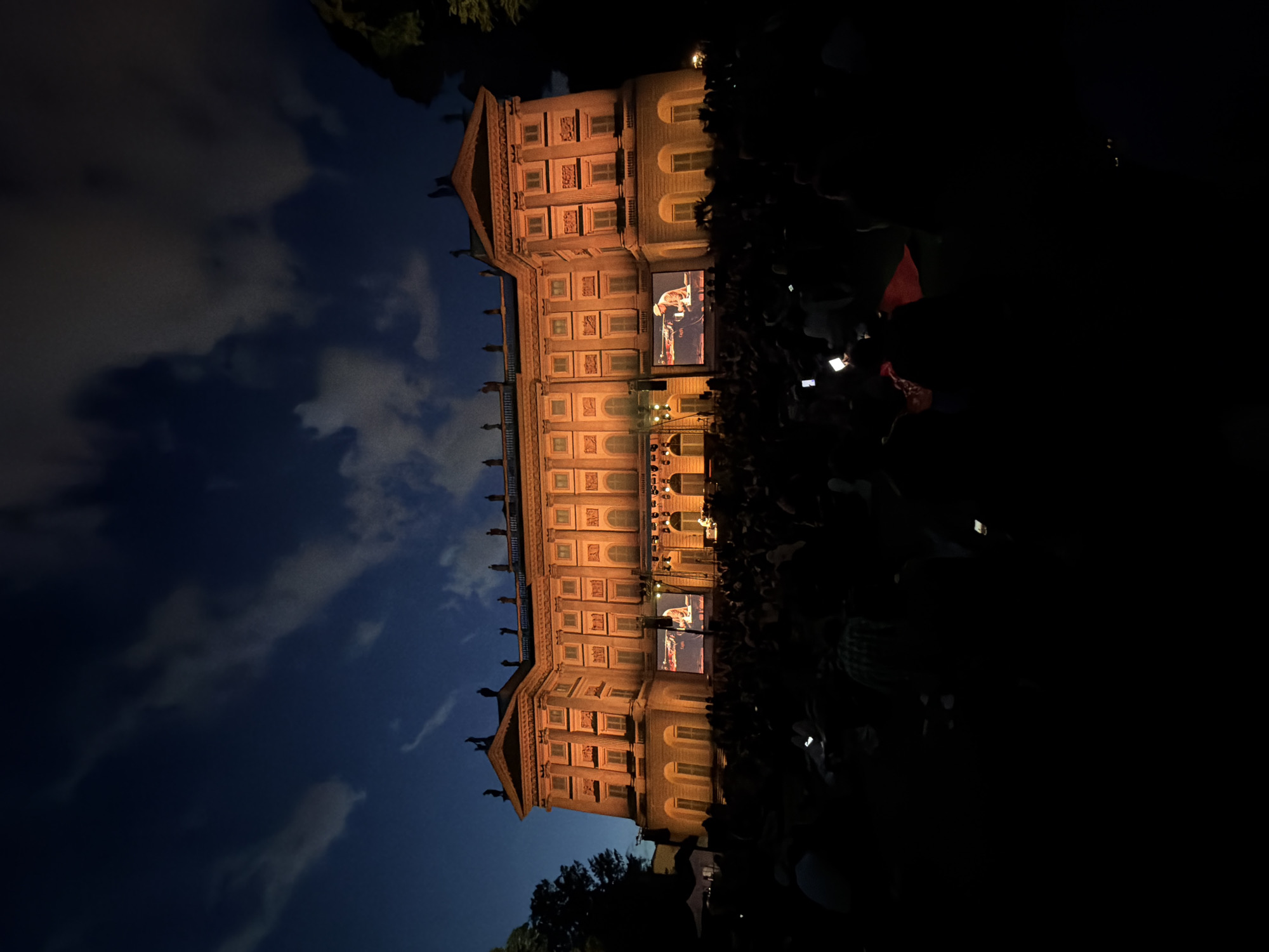 At night, a three-story building lit in warm light stands before a crowd. The facade features symmetrical rows of windows and decorative elements. Two large screens mounted on the building’s exterior display moving images. The sky above is a deep blue with scattered clouds. The darkness of the night surrounds the illuminated building, with some trees and vegetation visible at the bottom. The building is located in Milan, Italy.