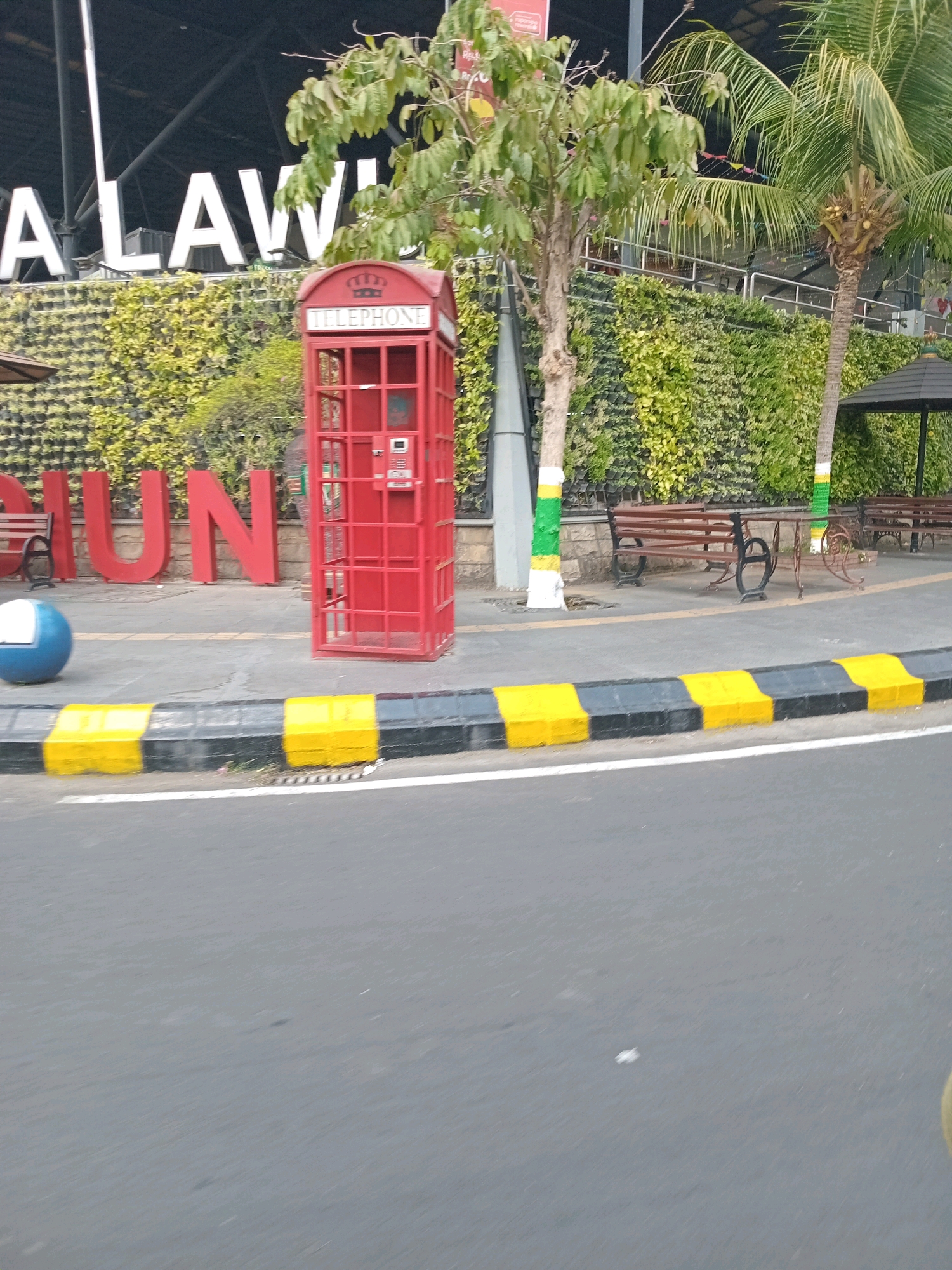 The image depicts an outdoor scene in an urban environment, featuring a red telephone booth, a green wall with a textured pattern, and large red letters spelling out "GIUN". The letters are positioned in front of the green wall, with some of them partially obscured by vegetation. A tree with sparse foliage stands to the left of the telephone booth, and a palm tree with coconuts is visible to the right. Several brown wooden benches with ornate black metal legs are placed along the walkway behind the telephone booth and green wall. A blue and white spherical object is located on the sidewalk to the left of the red letters. The ground in the foreground is an asphalt road with yellow and black striped markings along the curb. The background shows a modern building structure with a dark roof and exposed beams. The overall lighting suggests it is daytime.