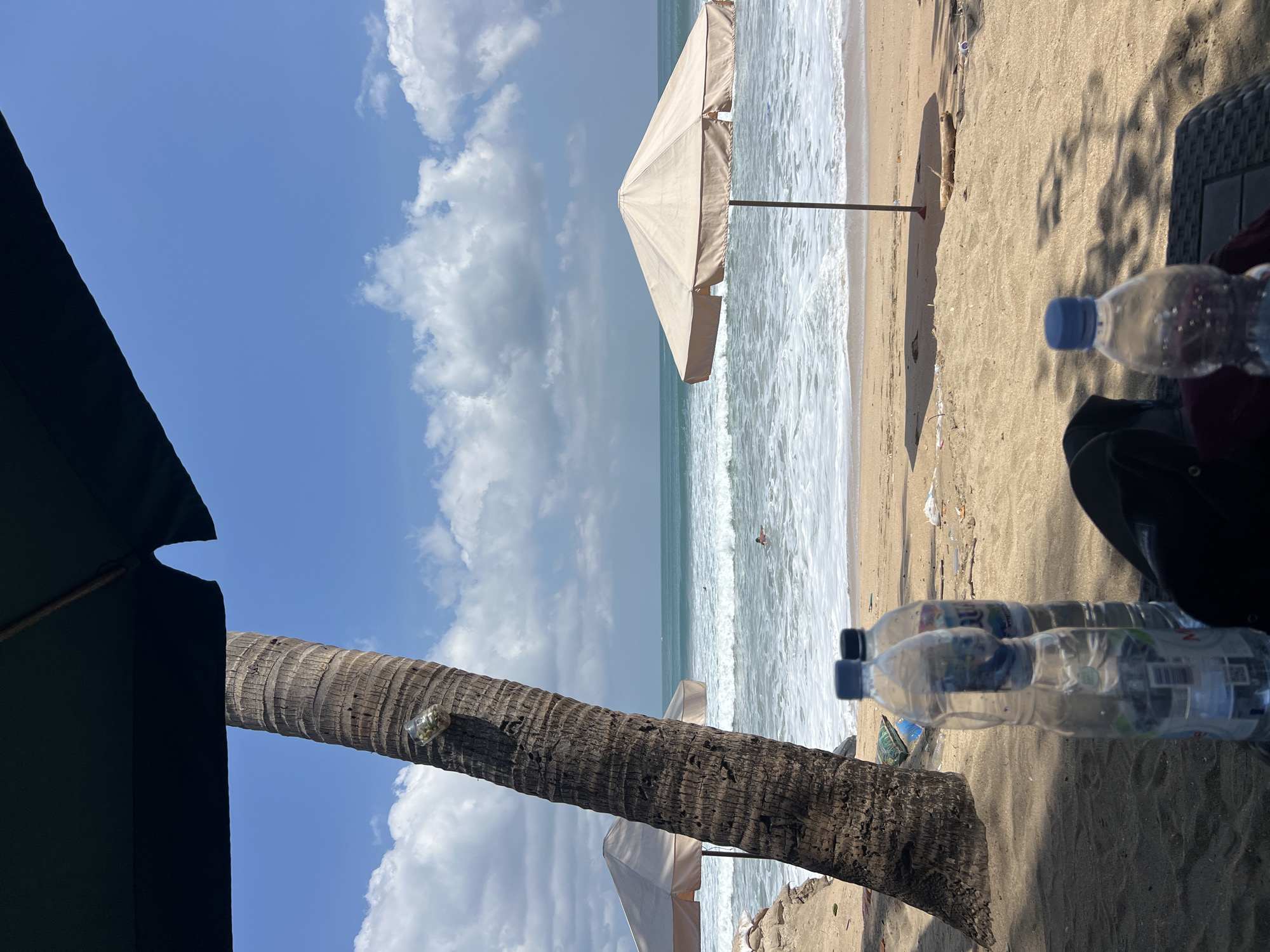 A wide-angle, eye-level shot captures a serene beach scene in Kuta, Indonesia, on a bright, partly cloudy day. In the foreground, partially obscuring the view, are the tops of two dark umbrellas. To the left, a thick, textured palm tree trunk rises, with a small glass jar containing what appears to be small stones or shells attached to its upper section. In front of the palm tree, two clear plastic water bottles stand upright on the sandy ground.
The middle ground of the image reveals a sandy beach where several white beach umbrellas are set up. One prominent, light-colored umbrella stands centrally on the right, casting a shadow on the sand. Further back, another similar umbrella is visible to the left, partially behind the palm tree. The ocean stretches out beyond the beach, with gentle waves breaking and creating white foam as they approach the shore. Two figures are discernible in the water; one is swimming, and another appears to be floating or wading.
The background is dominated by a vibrant blue sky adorned with scattered, fluffy white clouds. The sand on the beach shows signs of being recently disturbed, with some scattered debris, including small pieces of wood and plastic. The overall impression is one of a peaceful, sunny day at a tropical beach resort.