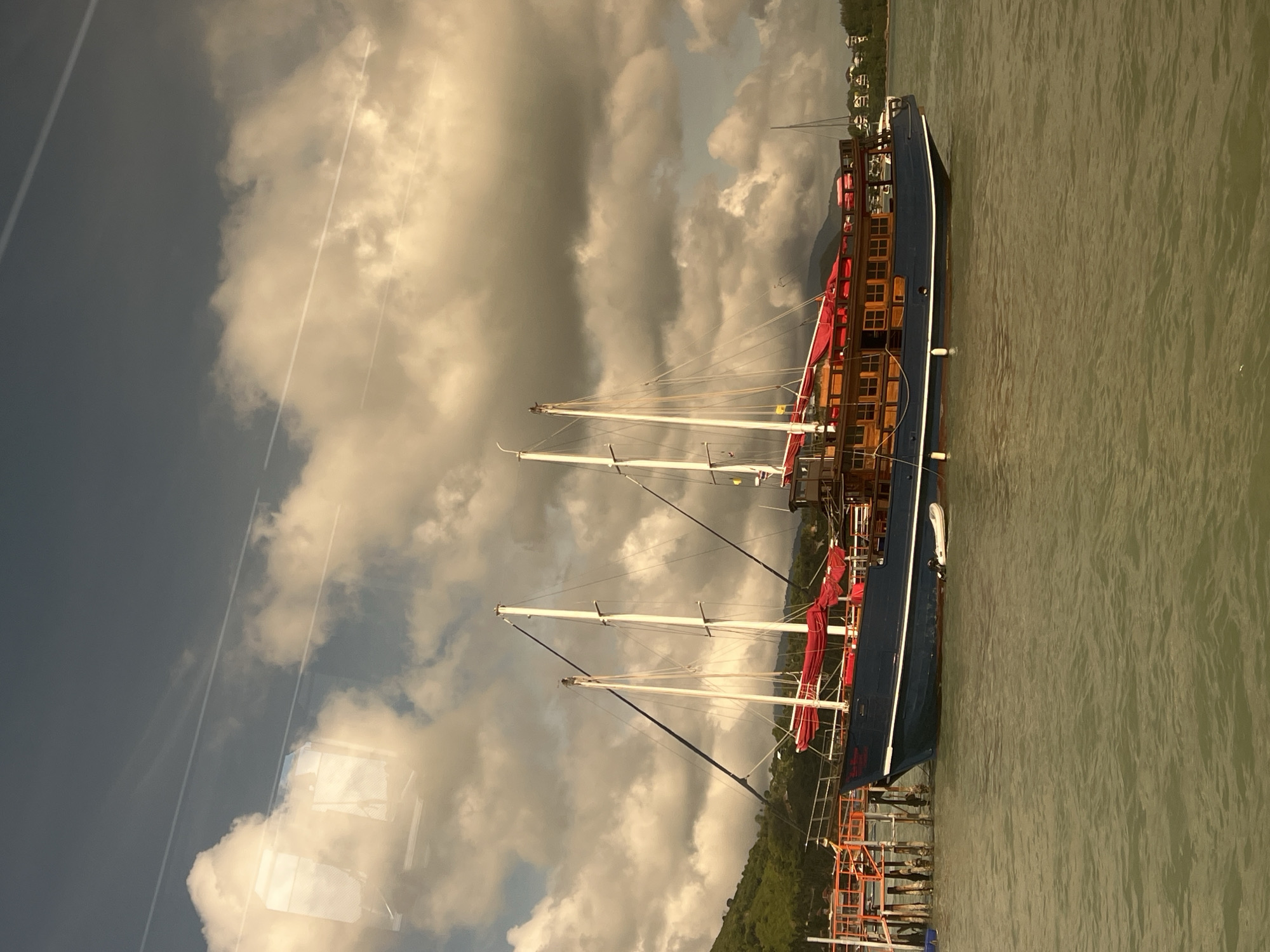 A large, blue wooden boat with three masts is docked at a pier in Baan Bang Rak, Thailand. The boat has red sails and a wooden upper deck with many windows. A small, white dinghy is tied to the side of the boat. The water is a murky green and appears choppy. In the background, there are green, tree-covered hills and a few buildings. The sky is cloudy with some sunlight breaking through. The image appears to have been taken through a window, as evidenced by reflections in the upper left portion of the image.