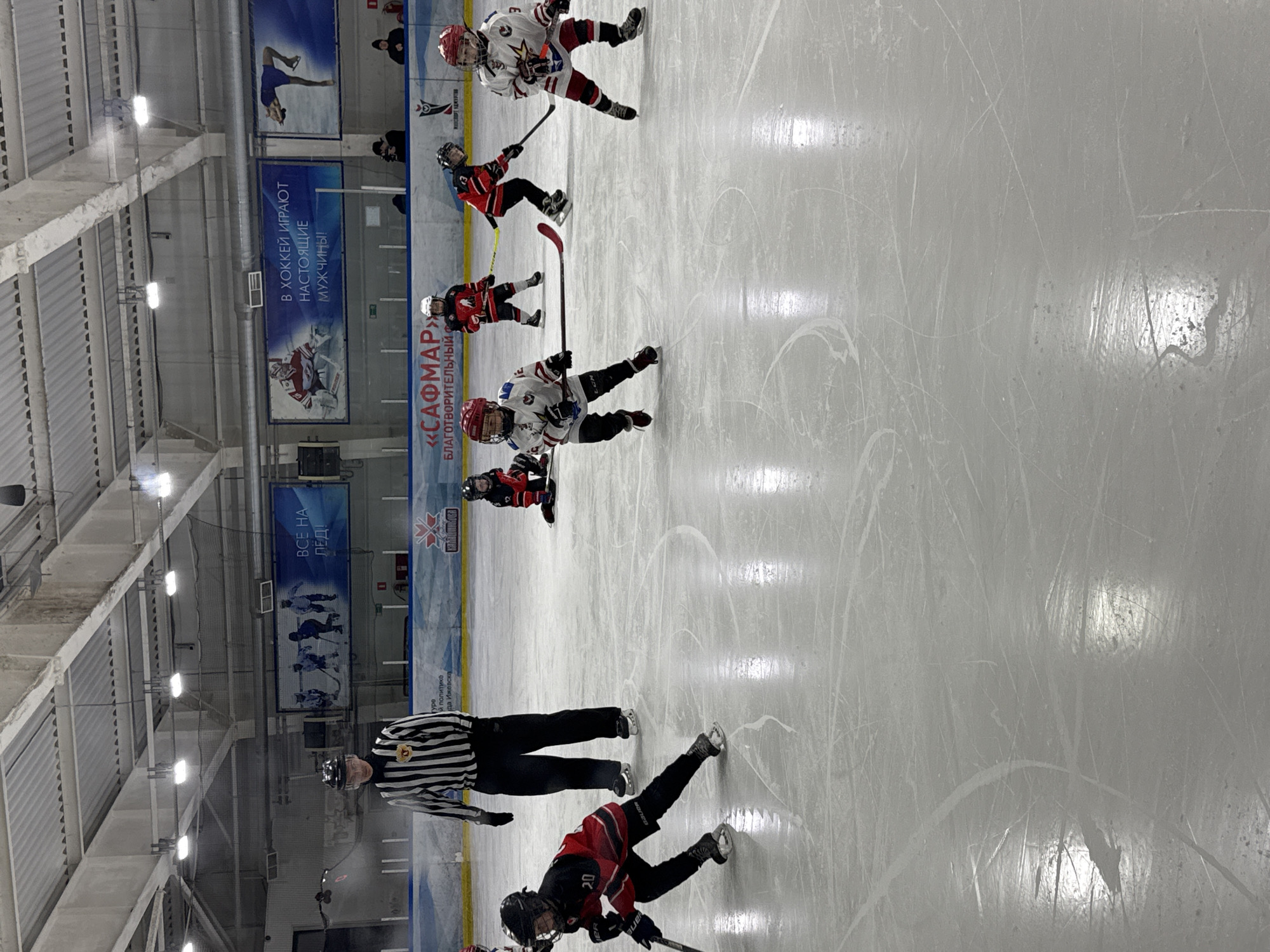 The image shows a hockey game in progress, likely involving young players. Several skaters are on the ice, wearing red and black uniforms, and white and red uniforms. A referee in a black and white striped uniform is also visible. The ice rink is surrounded by boards and advertisements. Banners on the wall in the background read "ВСЕ НА ЛЕД!" and "В ХОККЕЙ ИГРАЮТ НАСТОЯЩИЕ МУЖЧИНЫ!" Another banner includes the words "САФМАР", "БЛАГОТВОРИТЕЛЬНЫЙ", and "МАНСПОРТ УДМУРТИ". The text "Удмуртской политко" and "да Ижевска" indicates this is in the city of Izhevsk, Russia.