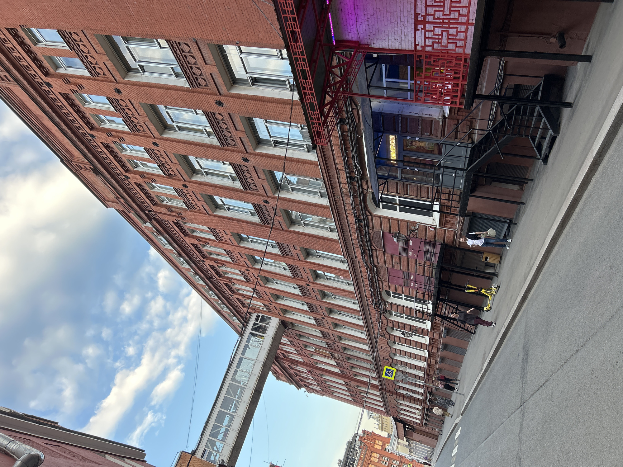 The image captures an urban scene featuring a tall, red brick building with numerous windows, displaying a repetitive pattern. A skyway bridge connects different sections of the building at a higher level. The street-level facade of the building features staircases, signage, and decorative elements, suggesting a commercial or mixed-use function. Pedestrians are visible on the sidewalk, with some individuals appearing to be in motion, walking or using scooters. The street itself is a gray paved surface, suggesting a typical urban thoroughfare. The sky above shows a mix of blue and white clouds, contributing to the natural light in the scene. The architecture of the building, along with the street-level activities, points to a location within a city, though the specific city is not identifiable from the image.