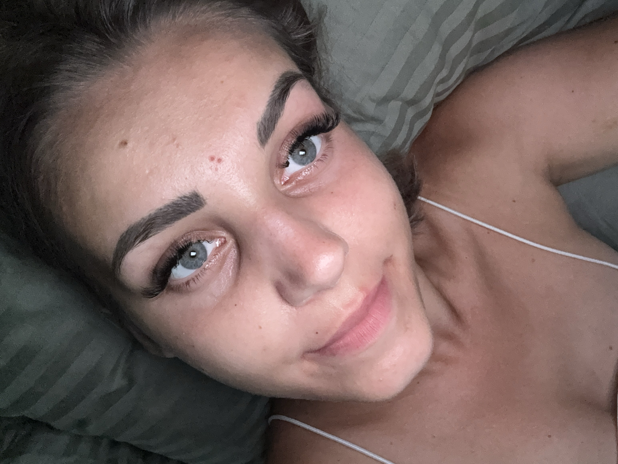 The image shows a close-up, overhead view of a woman lying down, likely in bed, with her head resting on a green striped pillow. She has light blue eyes, dark eyebrows, and what appear to be eyelash extensions. Her mouth is slightly open, revealing pink lips, and she has a mole on her cheek and a small mark on her forehead. She is wearing a white spaghetti strap top. The lighting suggests it is indoors, possibly at night or in a dimly lit room. The image does not contain any visual information that would indicate its location in Yuzhno-Sakhalinsk, Russia.