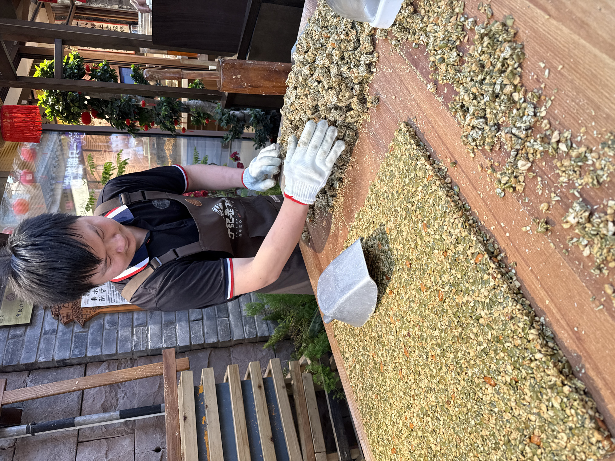 An overhead, slightly angled shot captures a young person meticulously preparing a traditional food product on a large wooden surface in what appears to be an outdoor or semi-outdoor marketplace. The person, likely male, wears a black polo shirt with striped trim, a brown apron featuring Chinese text ("YUJI ANCIE" and "百年匠心" - "century-old craftsmanship"), and white gloves with red cuffs. Their gloved hands are actively pressing and shaping a large mound of a granular mixture, which consists predominantly of green seeds (likely pumpkin seeds), light-colored nuts, and other coarse ingredients, possibly destined to become snack bars or candy. A metal scoop rests nearby on the wooden surface, and a section of the already flattened mixture is visible.
The background reveals elements of traditional Chinese architecture, including brick walls, wooden beams, and glass shopfronts. Decorative plants adorned with red ornaments or small lanterns add to the lively atmosphere. The bright sunlight suggests a clear day. Various Chinese characters are visible on signs and plaques, including "十八梯上老招牌" (Old brand on Shibati), which provides a strong clue to the location being Shibati, a historical area in Yuzhong District, China. Other visible text includes "市级非", "水香", "井滋", and "丁記者" (Reporter Ding), hinting at local recognition or brand details. The scene depicts a focused individual engaged in artisanal food preparation.