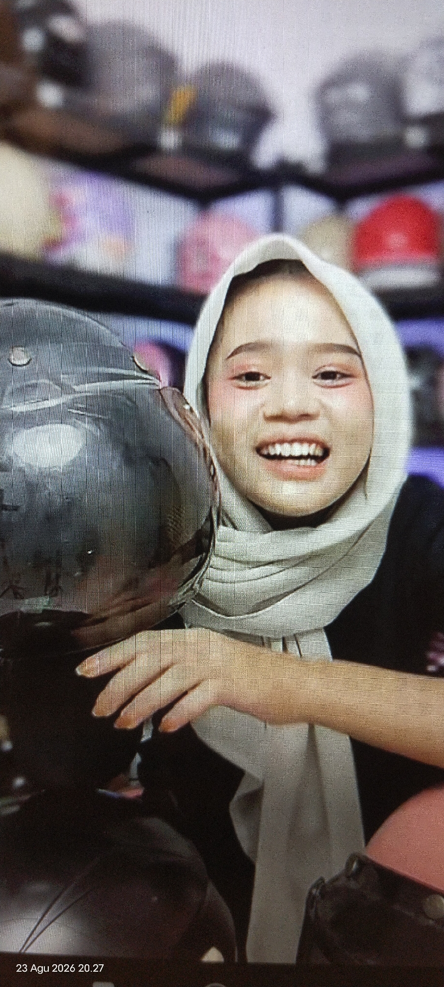 In East Jakarta, Indonesia, a young woman wearing a light-colored hijab and dark clothing is captured in a close-up shot. She is smiling broadly, with her mouth open and teeth visible. Her cheeks are adorned with prominent blush, and her eyes are lined with makeup. To her left, a portion of a dark, reflective object, possibly a helmet, is visible. The background is blurred, but appears to be a retail environment with shelves containing various items, some of which are colored in shades of purple and red, suggestive of caps or hats. A timestamp at the bottom left of the image reads "23 Agu 2026 20.27."