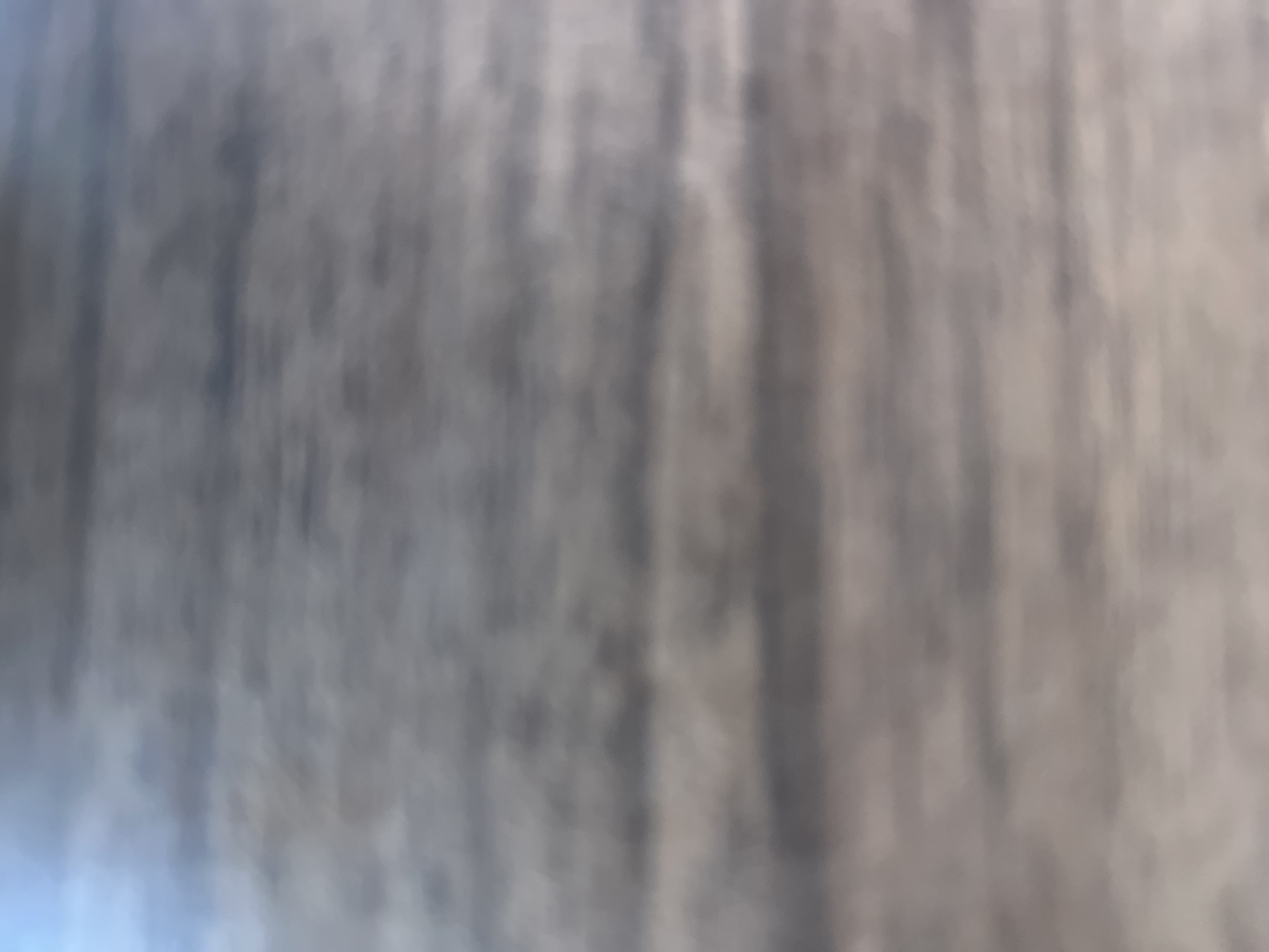 The image displays an extremely blurred and out-of-focus scene, contextually identified as being located in the city of Casablanca, Morocco. The visual content is abstract, characterized by soft, horizontal bands of varying color and luminosity. The upper-left region exhibits a light blue and cool grey tone, transitioning into broader horizontal stripes of muted greys, browns, and off-white shades across the mid and lower sections. Darker, indistinct bands are interspersed among lighter, diffuse areas, creating a striped, ethereal quality. No discernible people, subjects, objects, or distinct environmental features are identifiable within the frame due to the pervasive blur.