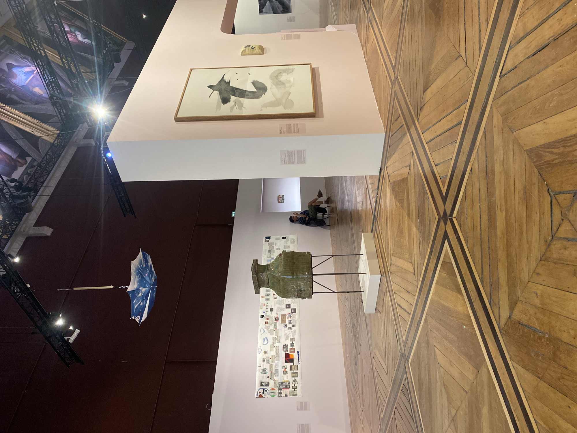 The image shows the interior of a building in Paris, France, likely a museum or art gallery. The floor is made of parquet wood. The walls are a light pink color. Hanging from the ceiling on the left side is an open, upside-down blue and white umbrella. A person is sitting in the background. There are several art pieces on display, including a framed ink drawing, a large green vase-like sculpture, and numerous informational posters.