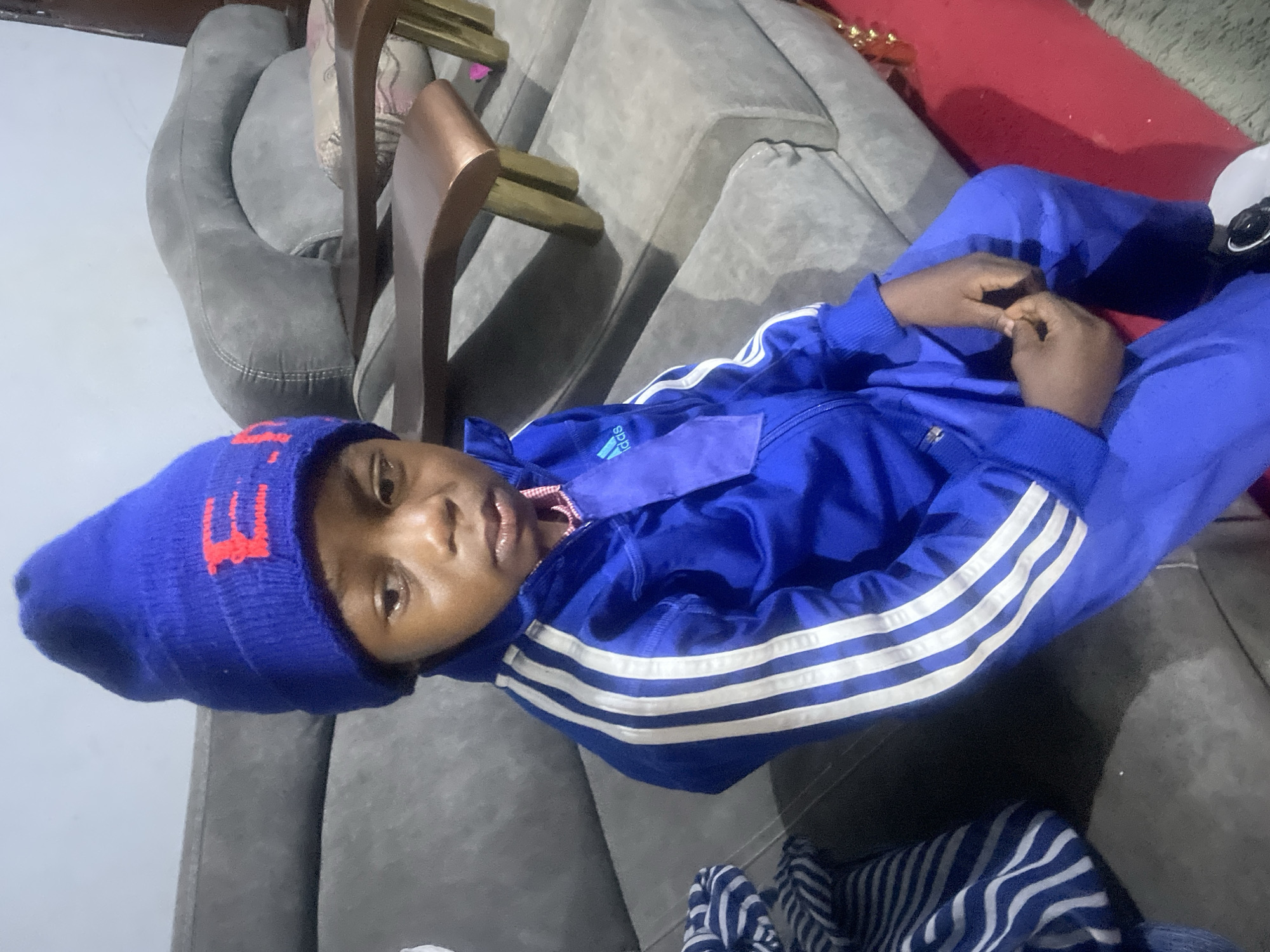A person, possibly a child, is lying on a gray sofa in what appears to be an indoor setting. The person is wearing a blue beanie with a red logo, a blue Adidas tracksuit, and white socks. The person is lying on their back with their legs crossed and their hands clasped together. The background includes sections of a gray sofa, parts of wooden chairs, and a red surface. The location is unknown and can't be determined based solely on the image. It is not possible to determine if the location is in Jos, Nigeria or any other specific city.