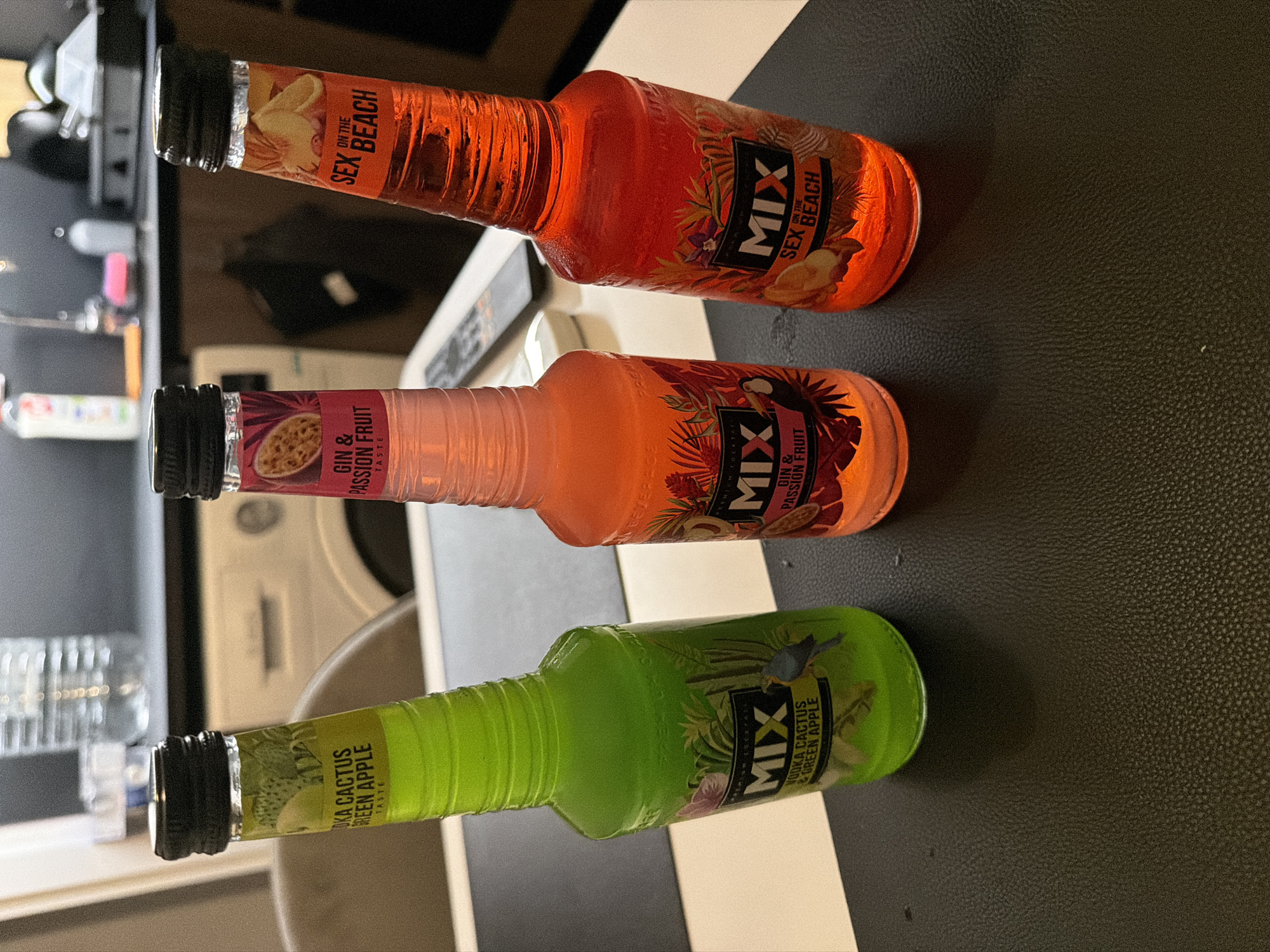 The image shows three bottles of "MIX" brand pre-mixed cocktails. From top to bottom, the flavors are "Sex on the Beach" (orange liquid), "Gin & Passion Fruit" (pinkish-orange liquid), and "Vodka Cactus & Green Apple" (green liquid). The bottles are arranged diagonally on a surface that appears to be dark leather. In the blurred background, there are hints of a kitchen setting, including appliances and glassware. While the "MIX" brand is sold in Plovdiv, Bulgaria, the location of this image cannot be definitively confirmed as Plovdiv based on visual information alone.