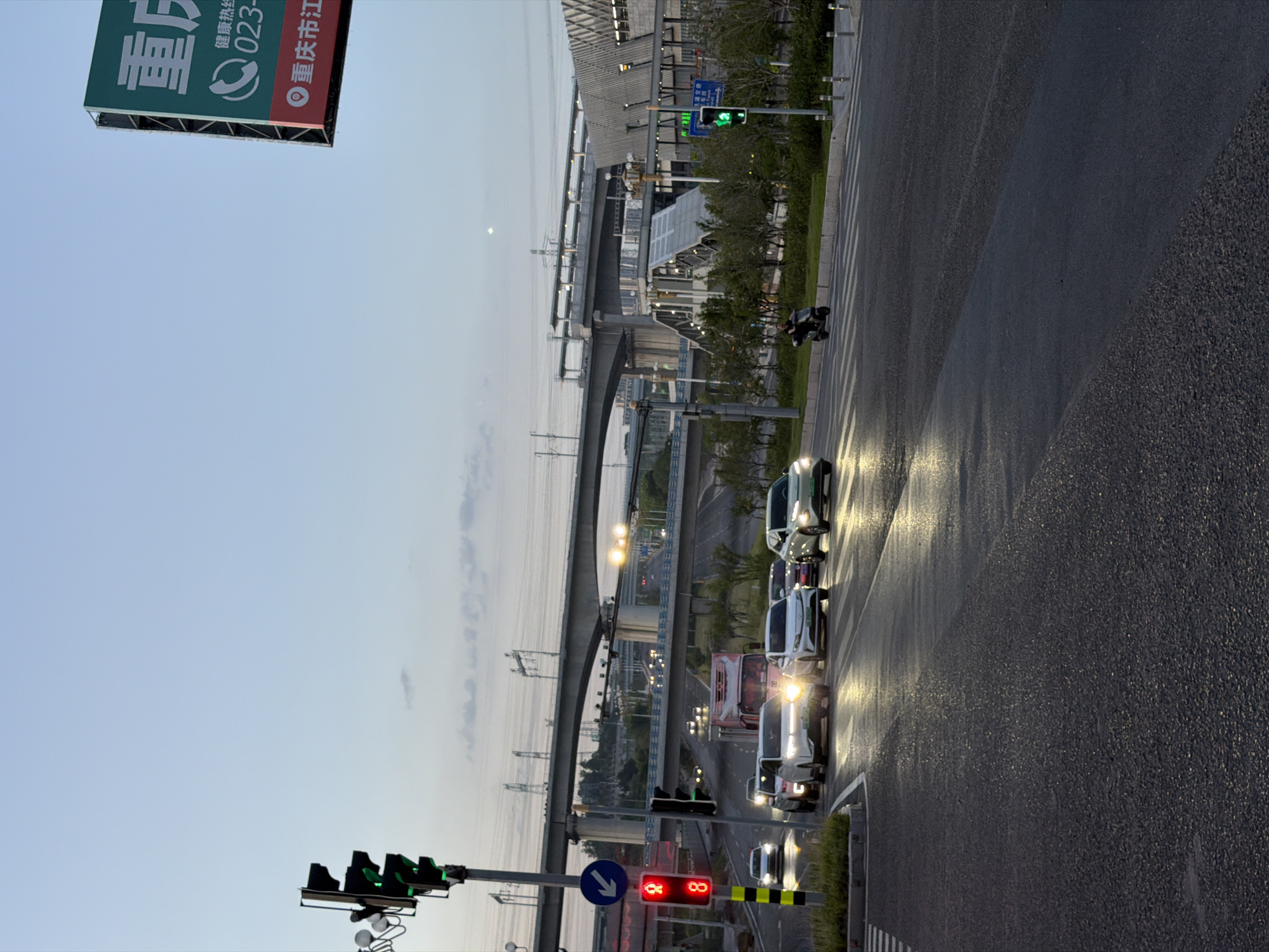 The image shows a street scene at dusk in Jiangjin, China. A traffic light with a red light and a countdown timer of '8' is visible on the left. Several cars are stopped at the intersection, waiting for the light to change. A truck is also visible further down the road. In the background, an elevated highway and a modern building are prominent. A scooter rider is crossing the street in the mid-ground. A large billboard with Chinese characters is positioned in the upper right corner. The sky is a pale blue with some scattered clouds. The road surface is wet, suggesting recent rain.