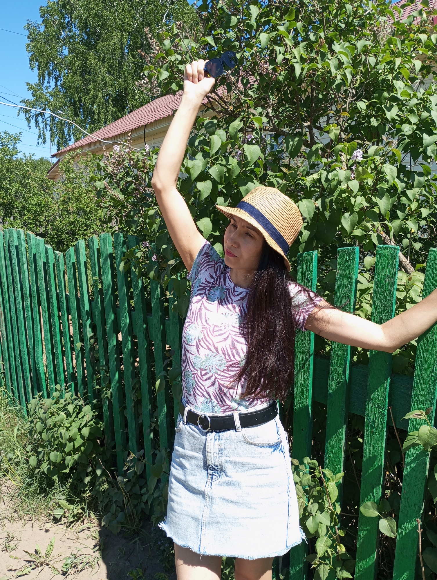 A woman stands beside a green wooden fence on a sunny day in Буренка, Russia. She wears a straw hat with a navy blue band, a patterned t-shirt, a denim skirt, and a black belt. Her dark hair falls down her back. She holds an object in her raised right hand. The background features a house with a reddish-brown roof, trees, and greenery.