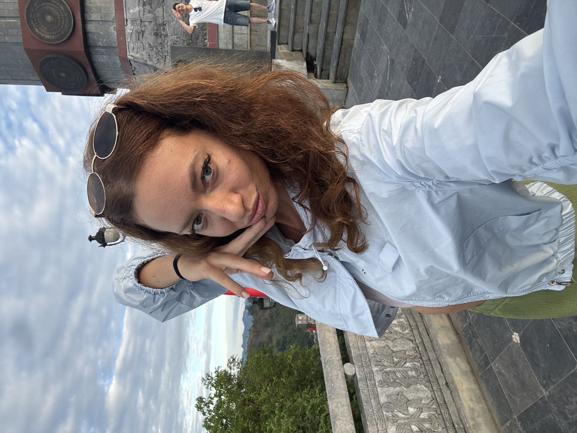 A woman poses for a selfie in Lũng Cú Commune, Vietnam. She has auburn curly hair, sunglasses perched on her head, and wears a light blue windbreaker. Her face is close to the camera, resting on her hand. In the background, a man stands on a set of stone steps, partially obscured. The environment features a cloudy sky, trees, and stone architectural elements.