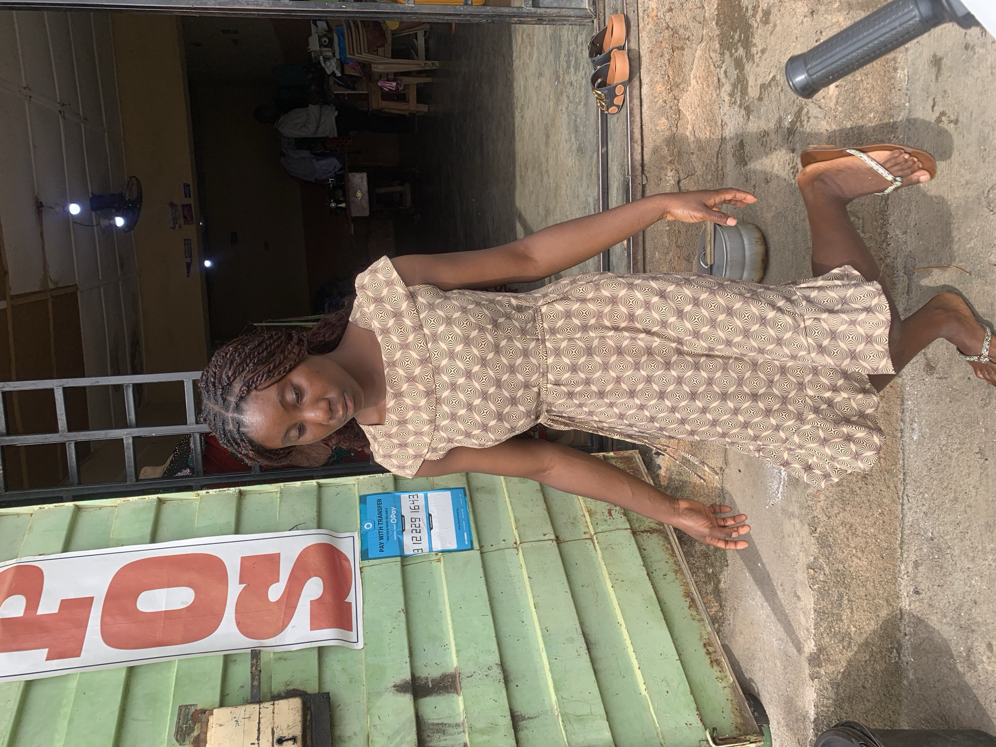 A young African woman with braided hair stands on concrete steps in front of a green corrugated metal building. She wears a short-sleeved, patterned dress. To her left, a vertical sign with red lettering reads "POS". A blue payment sticker is affixed to the building below the sign. Two pairs of flip-flops rest on the steps behind her. The building's doorway reveals an interior with dim lighting, where two men and a sewing machine are visible. The scene is set in the city of Jos, Nigeria.